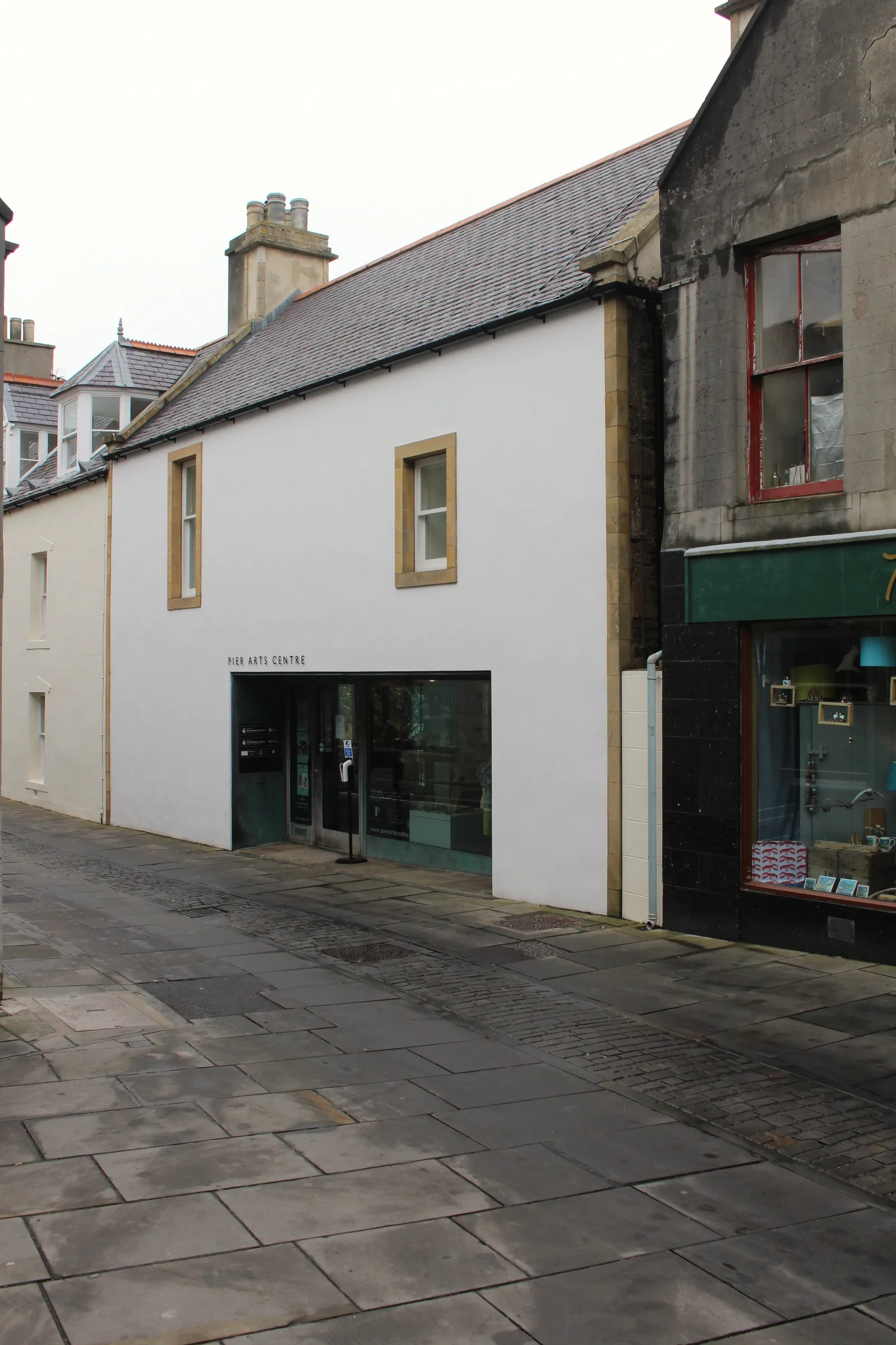 The street entrance of the Pier Arts Centre on Victoria Street, Stromness — a white-harled gable with the gallery name lettered above a glazed doorway, on the flagstone street