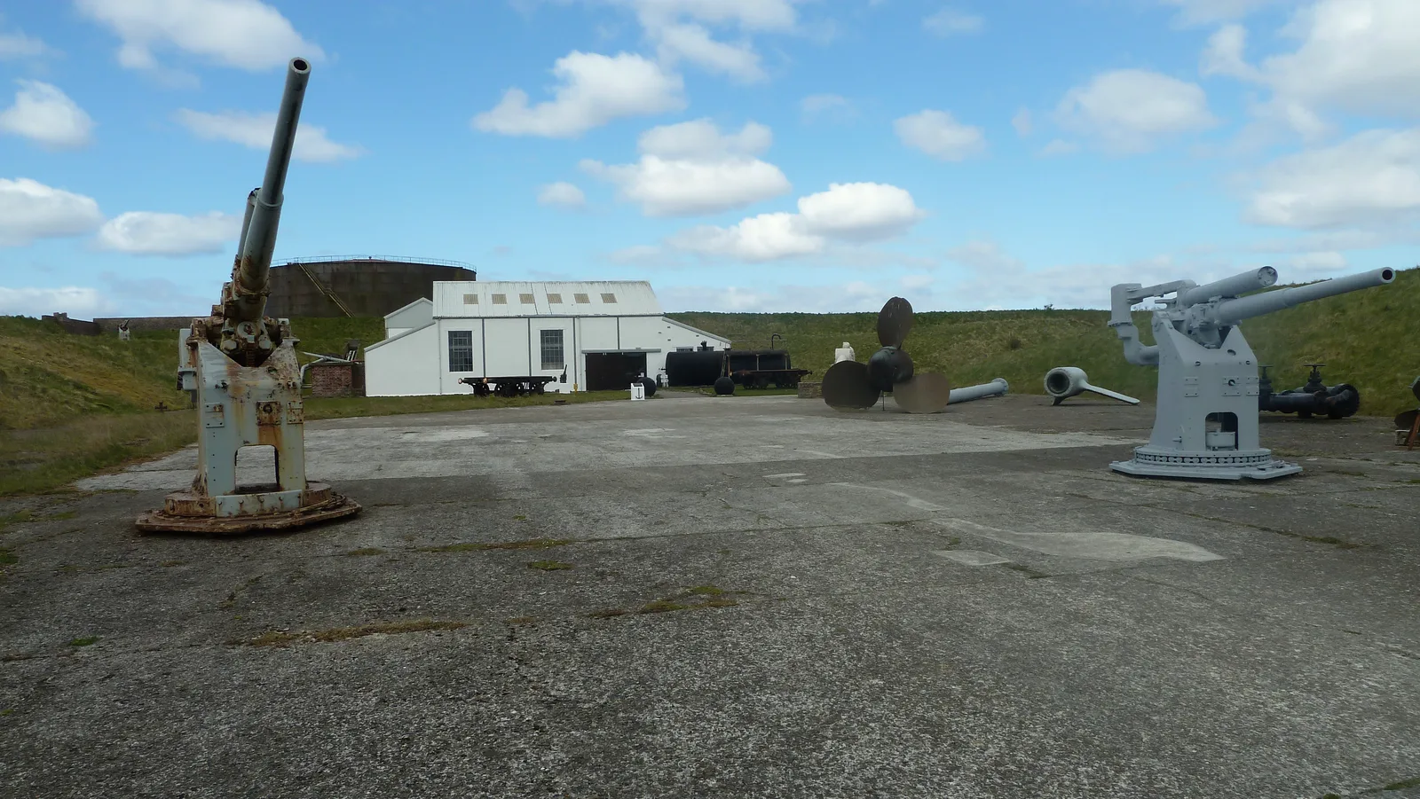 Open-air exhibits at the Scapa Flow Museum, Lyness, on the island of Hoy — two naval guns facing each other across a concrete apron, with a ship's propeller, a white-clad building and a fuel storage tank behind