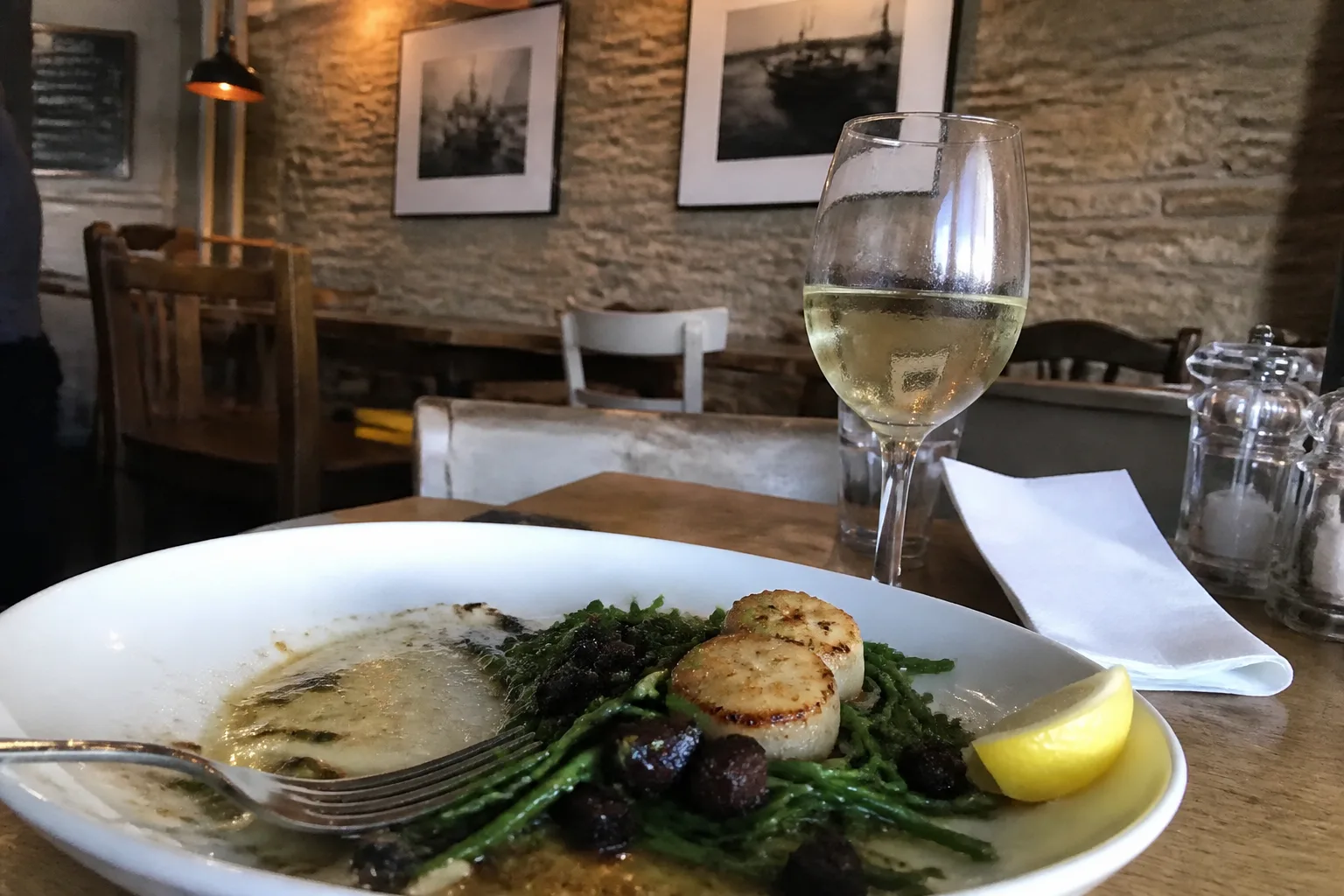 A small wooden restaurant table in Kirkwall, Orkney, with a half-eaten plate of pan-seared hand-dived Orkney king scallops on a bed of bright green samphire and small cubes of black pudding with a swirl of brown butter sauce, a glass of cold white wine with condensation, and warm pendant lamp light against an exposed sandstone wall in the background