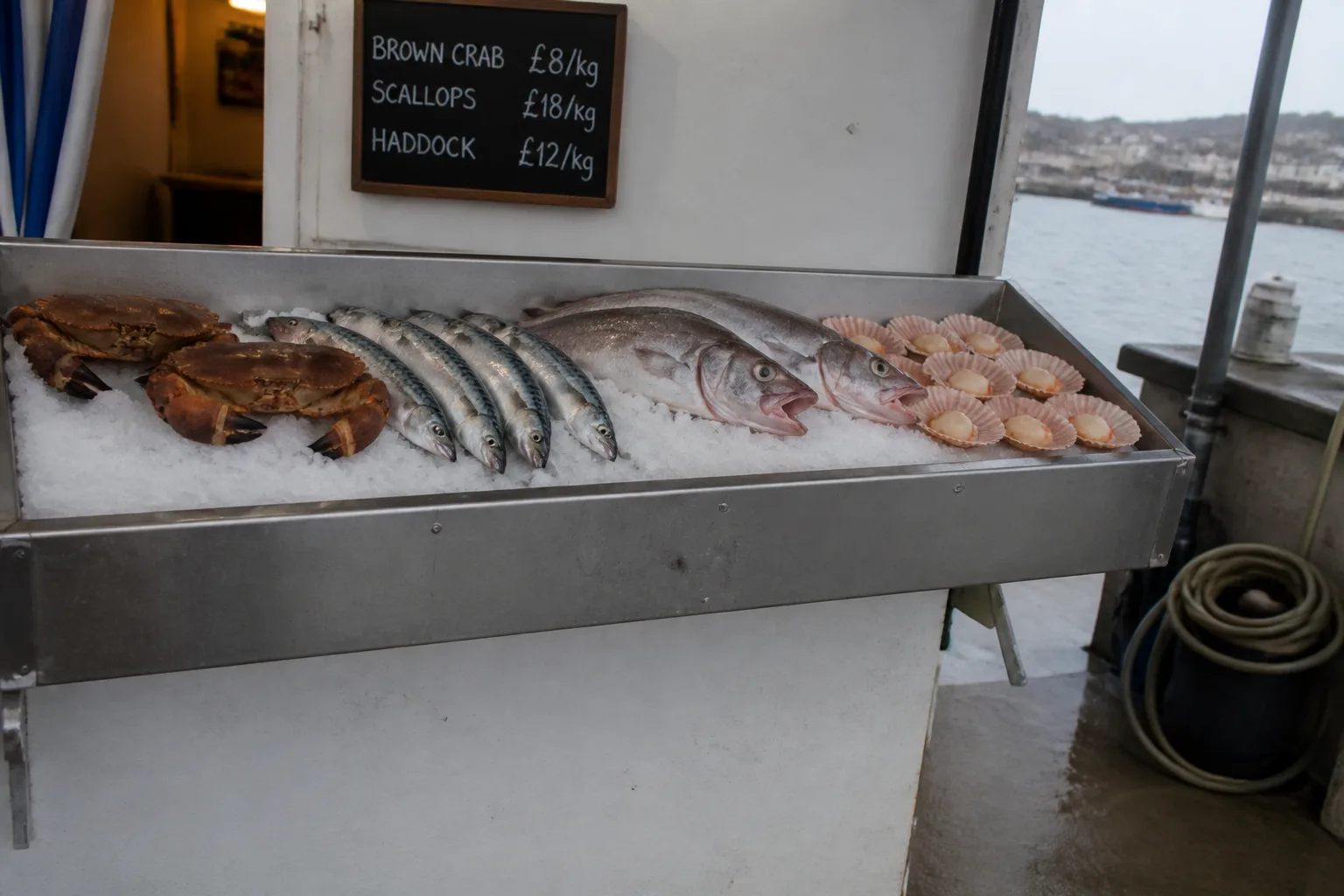 A small fishmonger's chilled display counter on the pier at Stromness harbour, Orkney, on a damp morning, with ice-bedded fresh fish — large brown crabs with claws bound, silver mackerel laid in a row, two whole haddock with pale pink gills, and a small heap of pale pink scallops in their shells — and a chalkboard listing the day's prices above