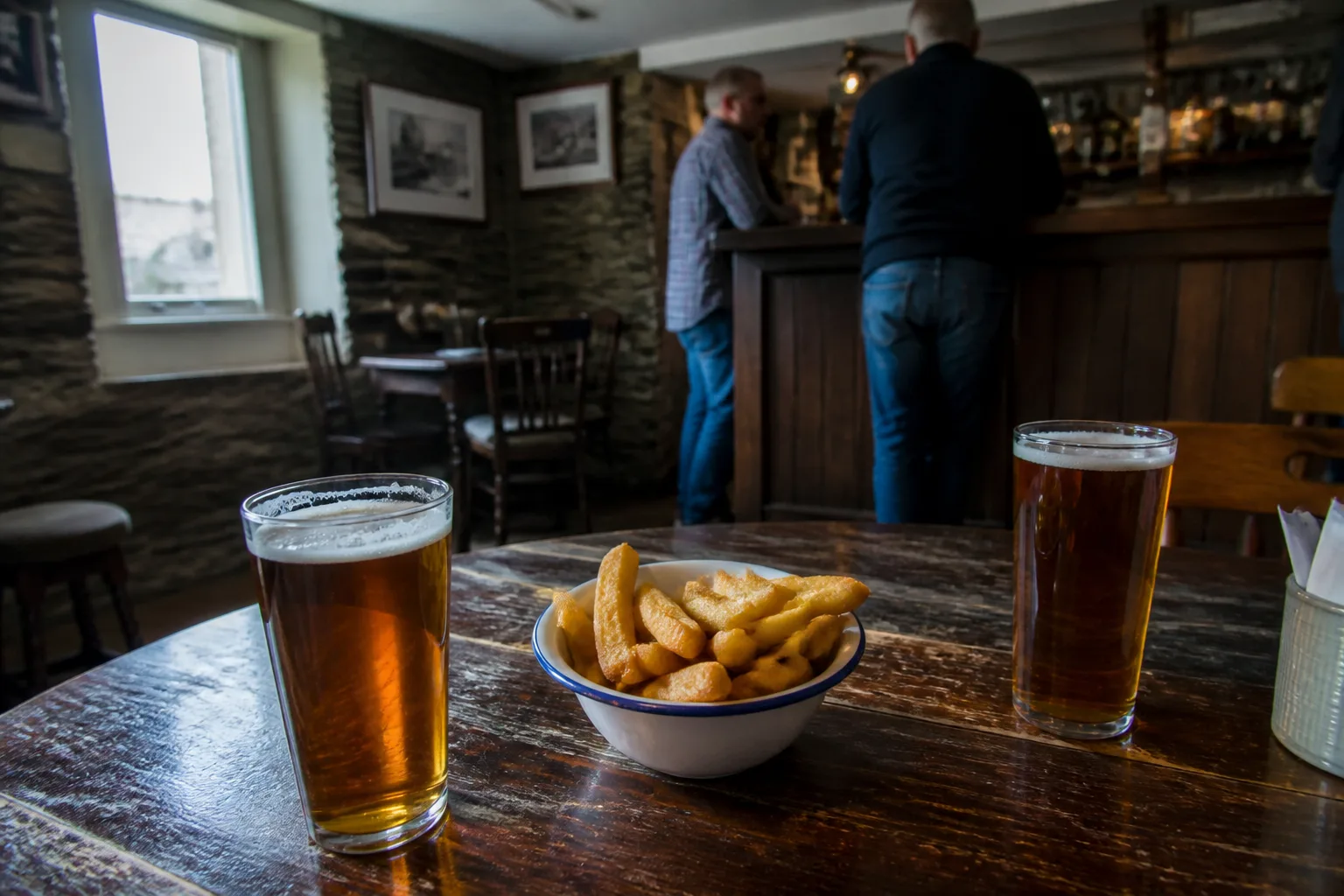Inside a cosy traditional Orkney pub at lunchtime — two pints of amber local ale and a bowl of chips on a worn dark-wood table, stone walls, daylight through a small window, locals at the bar