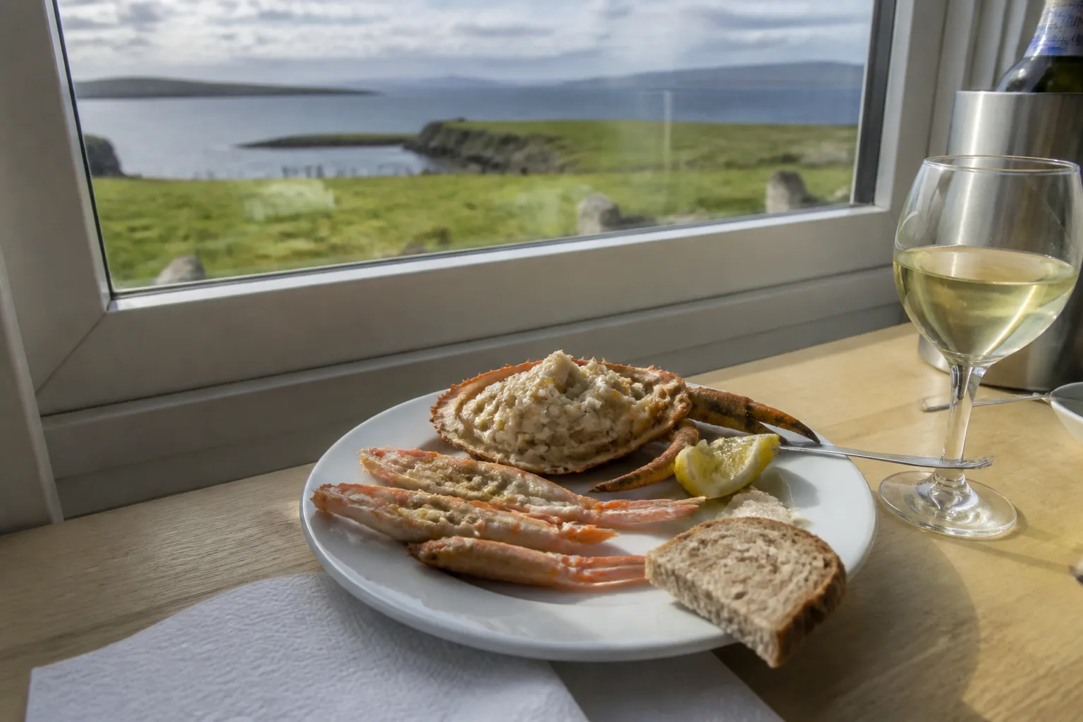 A plate of fresh Orkney seafood — half a dressed brown crab, langoustines and brown bread with a glass of white wine — on a restaurant table beside a window looking out over green Orkney coast and sea