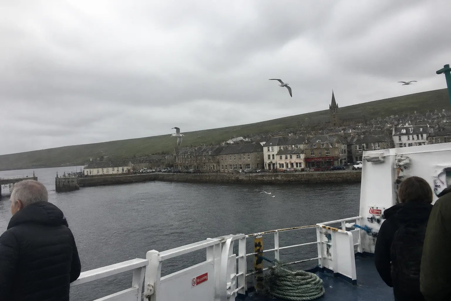 View from the rear deck of a NorthLink ferry approaching Stromness harbour, Orkney, on an overcast morning — slate-roofed sandstone houses lining the harbour with a church spire above, low green hills of West Mainland under heavy grey cloud, a few seagulls hovering over the white-painted ferry rail