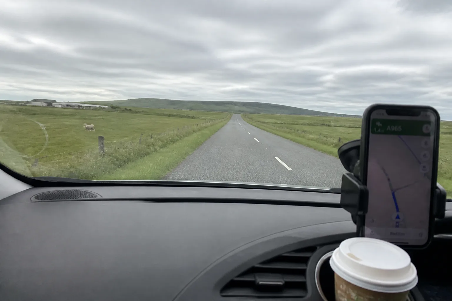 View from the windscreen of a small white hire car driving on the empty single-track A965 road across the West Mainland of Orkney, with a smartphone in a holder showing Google Maps and a paper coffee cup in the cupholder, sheep visible in the field and low hills under overcast sky