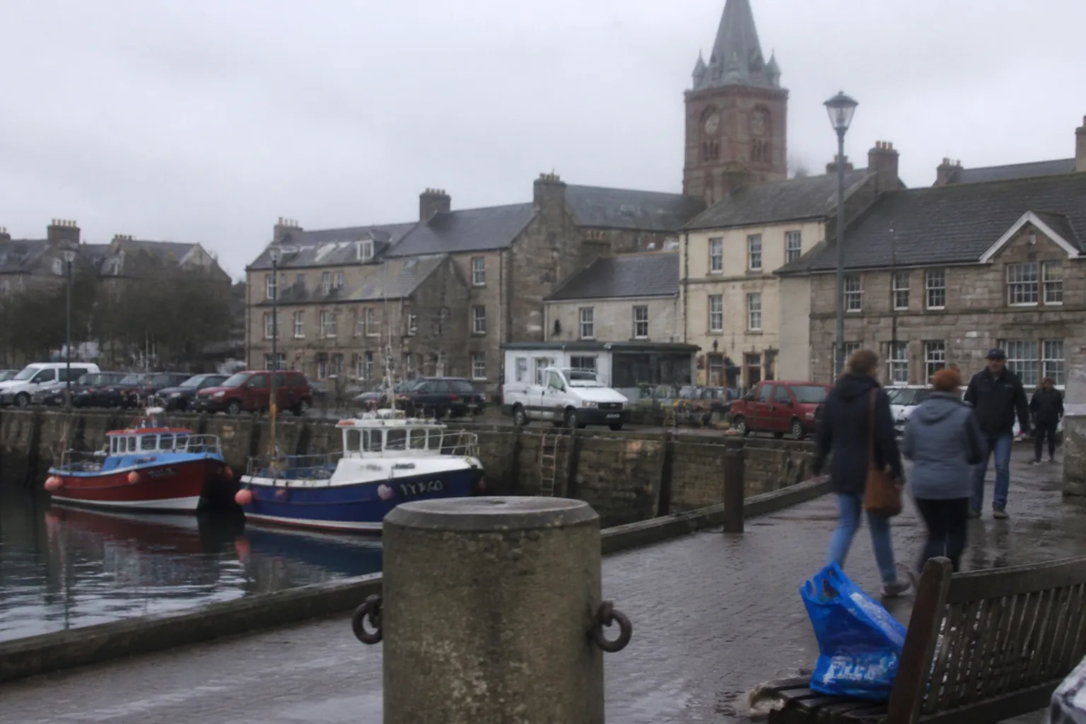 Kirkwall harbour, Orkney, on a mild morning with a few small fishing boats moored at the stone pier and the distinctive sandstone tower and copper-clad spire of St Magnus Cathedral visible above the rooftops in the background