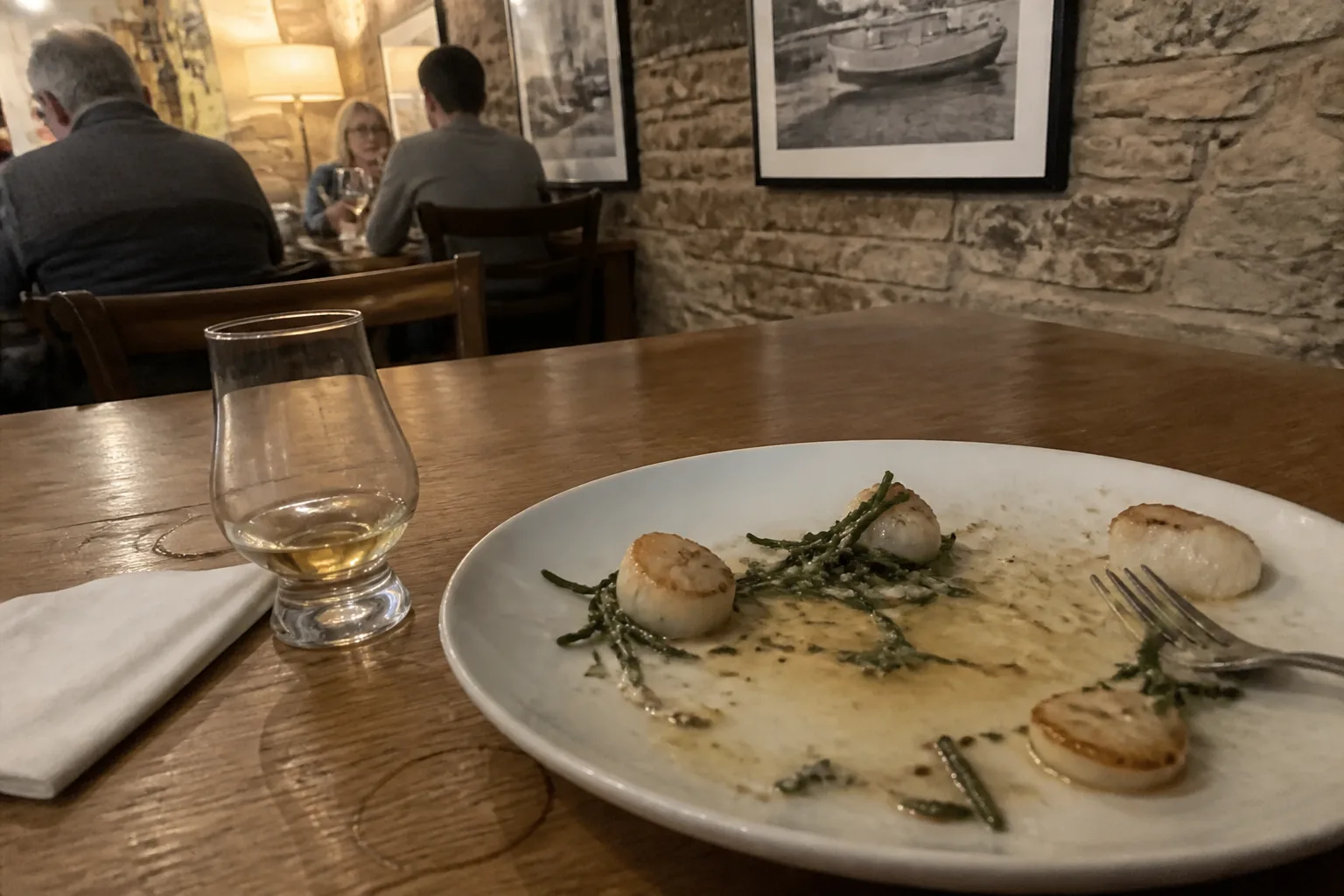 Small restaurant table in central Kirkwall, Orkney, in the evening with a half-eaten plate of locally-caught hand-dived scallops with samphire, a small glass of Scapa whisky, and a folded paper napkin on a wooden tabletop with warm lamp light