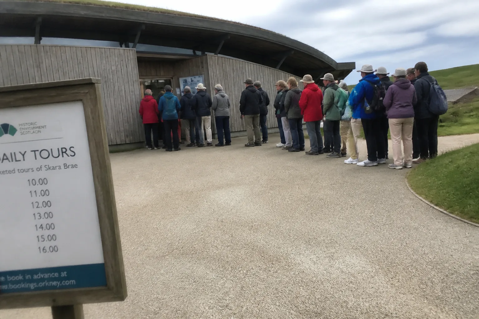 Modest queue of about a dozen visitors in waterproof jackets and sun hats waiting at the timber-clad ticket building of Skara Brae visitor centre, Orkney, on a busy summer afternoon