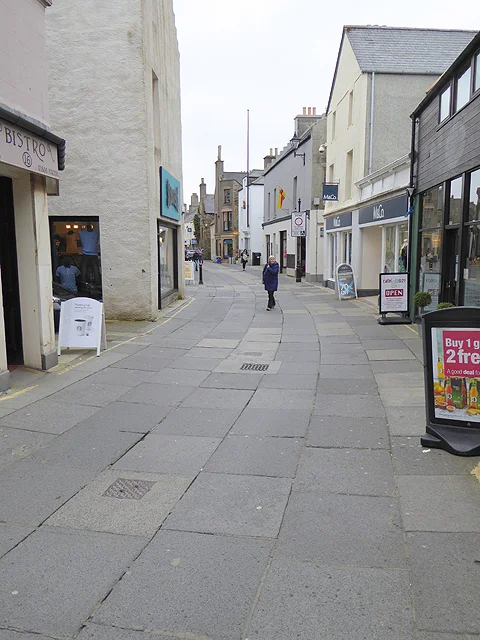 Albert Street, the main shopping street in central Kirkwall, Orkney, on a wet day — flagstone-paved pedestrian street lined with independent shopfronts, bunting strung overhead, a few shoppers in waterproofs, with the sandstone wall of St Magnus Cathedral precinct on the right