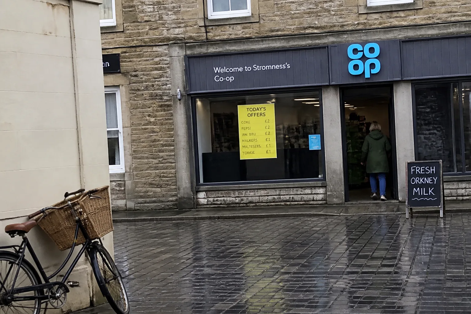 The exterior of the small Co-operative Food shop on a cobblestone street in central Stromness, Orkney — a sandstone shopfront with a blue Co-op logo, a printed yellow TODAY'S OFFERS poster taped inside the window, a chalkboard A-board on the cobbles reading FRESH ORKNEY MILK, a basket bicycle parked against the wall on the left and a woman in a green waterproof entering the shop