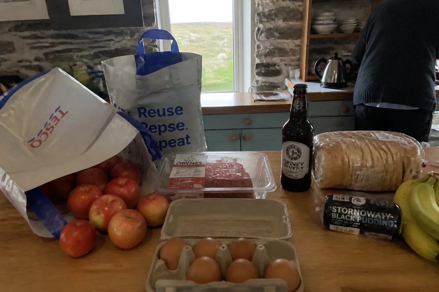 A small Orkney holiday-let kitchen with the morning's grocery shop just back from Tesco — two reusable Tesco carrier bags on the wooden countertop with one tipped over and apples spilling out, a plastic-wrapped pack of Orkney mince, a cardboard tray of six brown eggs, a bottle of Orkney Brewery beer, a sliced loaf of bread, a pack of black pudding, and a bunch of bananas, with a stone-walled kitchen window looking out to a sheep field behind