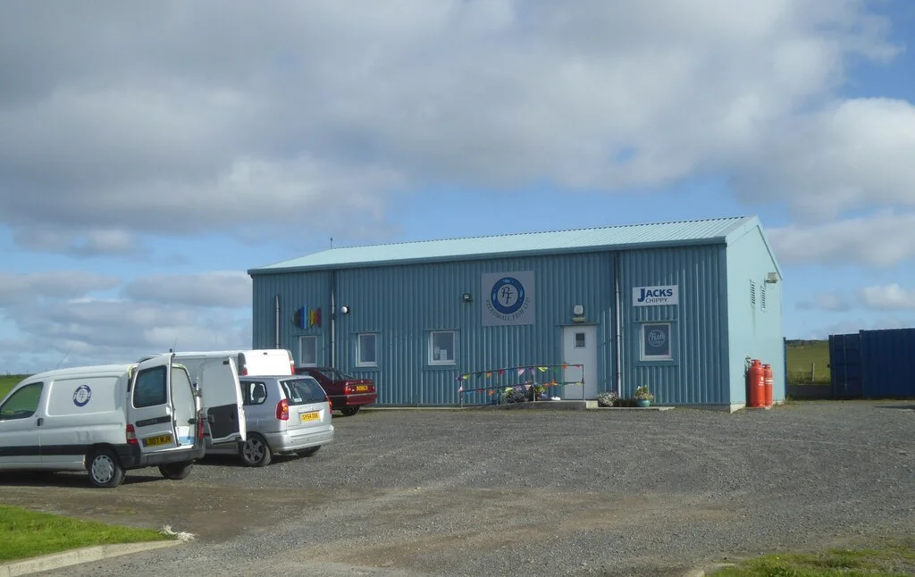 J C Tulloch general merchant shop and Post Office at Pierowall on the island of Westray, Orkney — a low harled building with a J C Tulloch GENERAL MERCHANTS sign and a Post Office sign, a red Royal Mail van and several cars parked outside under a bright sky