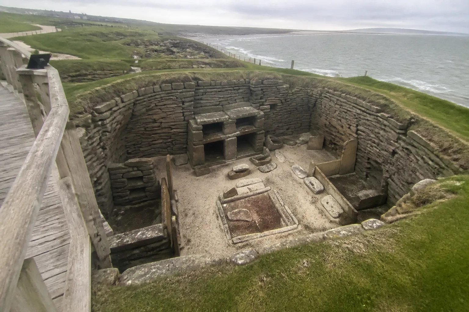 Skara Brae Neolithic stone house viewed from the modern wooden visitor walkway — thick drystone walls of dark grey flagstone enclosing a square room with a stone hearth in the middle, stone bed-boxes against the walls, and the famous stone dresser at the back, with the Bay of Skaill visible in the distance