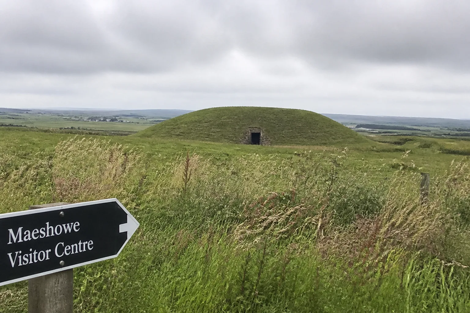 The exterior of Maeshowe Neolithic chambered tomb on Orkney Mainland — a large grass-covered earthen mound rising from flat farmland, with the dark rectangular stone entrance passage visible at its base and a directional sign pointing to the Maeshowe Visitor Centre on a wooden post in the foreground