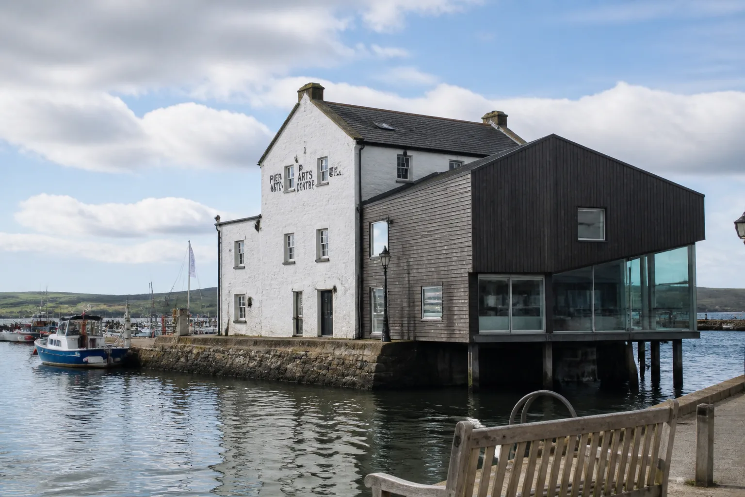 Environmental exterior photo of the Pier Arts Centre on Stromness pier in Orkney showing the contemporary architectural composition of a renovated 19th century stone pier house with whitewashed walls and slate roof joined to a strikingly modern dark-grey timber and glass extension that sits directly over the water of Stromness harbour with calm grey-blue sea in the foreground reflecting the building and a few moored small fishing boats off the pier under a partly-cloudy bright June sky