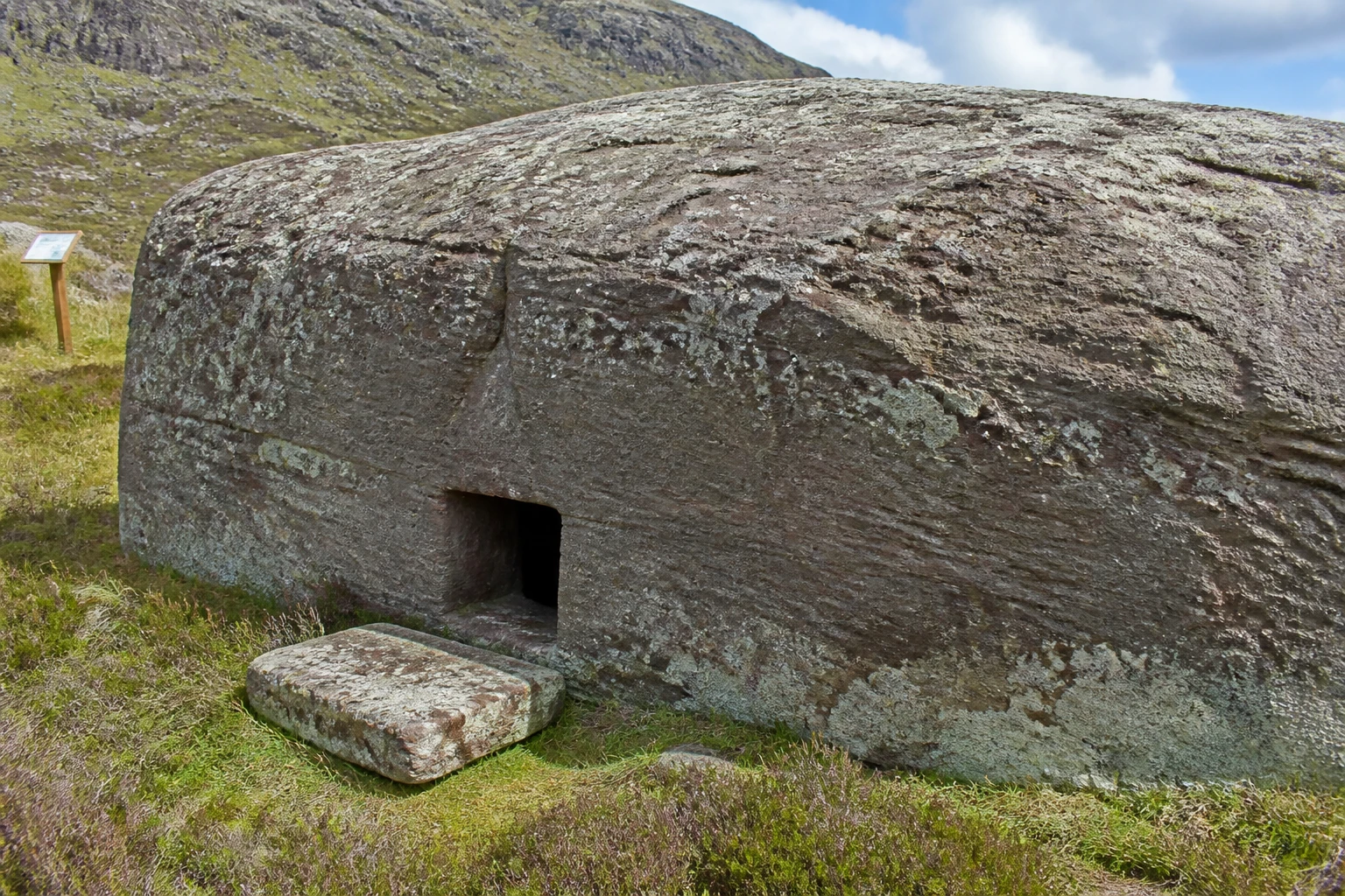 The Dwarfie Stane on Hoy Orkney — a single enormous block of weathered red-grey Old Red Sandstone half-buried in heathery moorland, with a small dark rectangular entrance carved into the side and a removable square sandstone plug-stone lying on the heather next to it, lichens visible on the rock surface