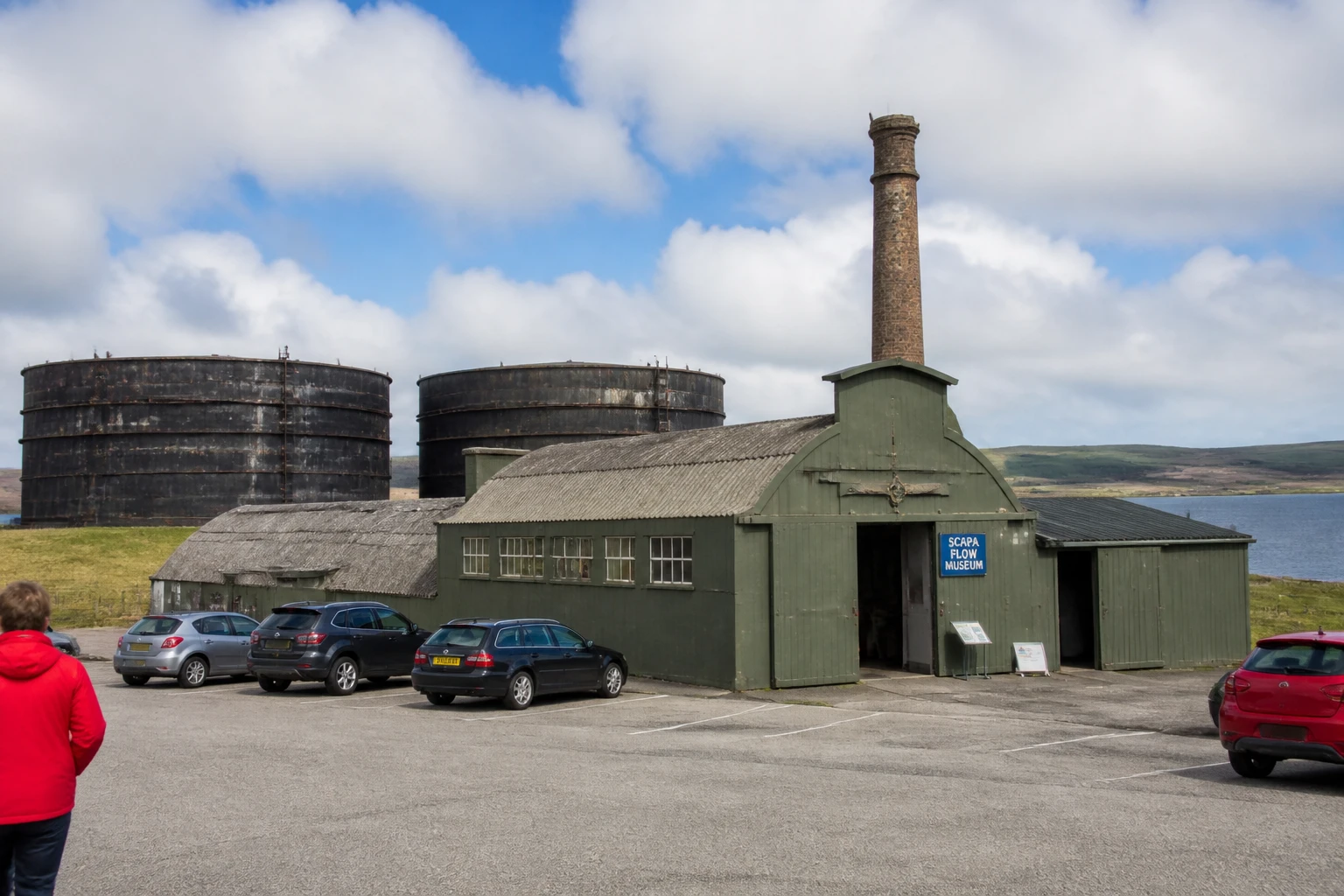 Scapa Flow Museum at Lyness on Hoy Orkney in a converted wartime pump-house and Romney hut painted muted dark green and grey with a tall brick chimney, two enormous circular naval oil storage tanks visible behind, modern signage panel reading SCAPA FLOW MUSEUM, tarmac car park in foreground, calm Scapa Flow water and Mainland hills in distance