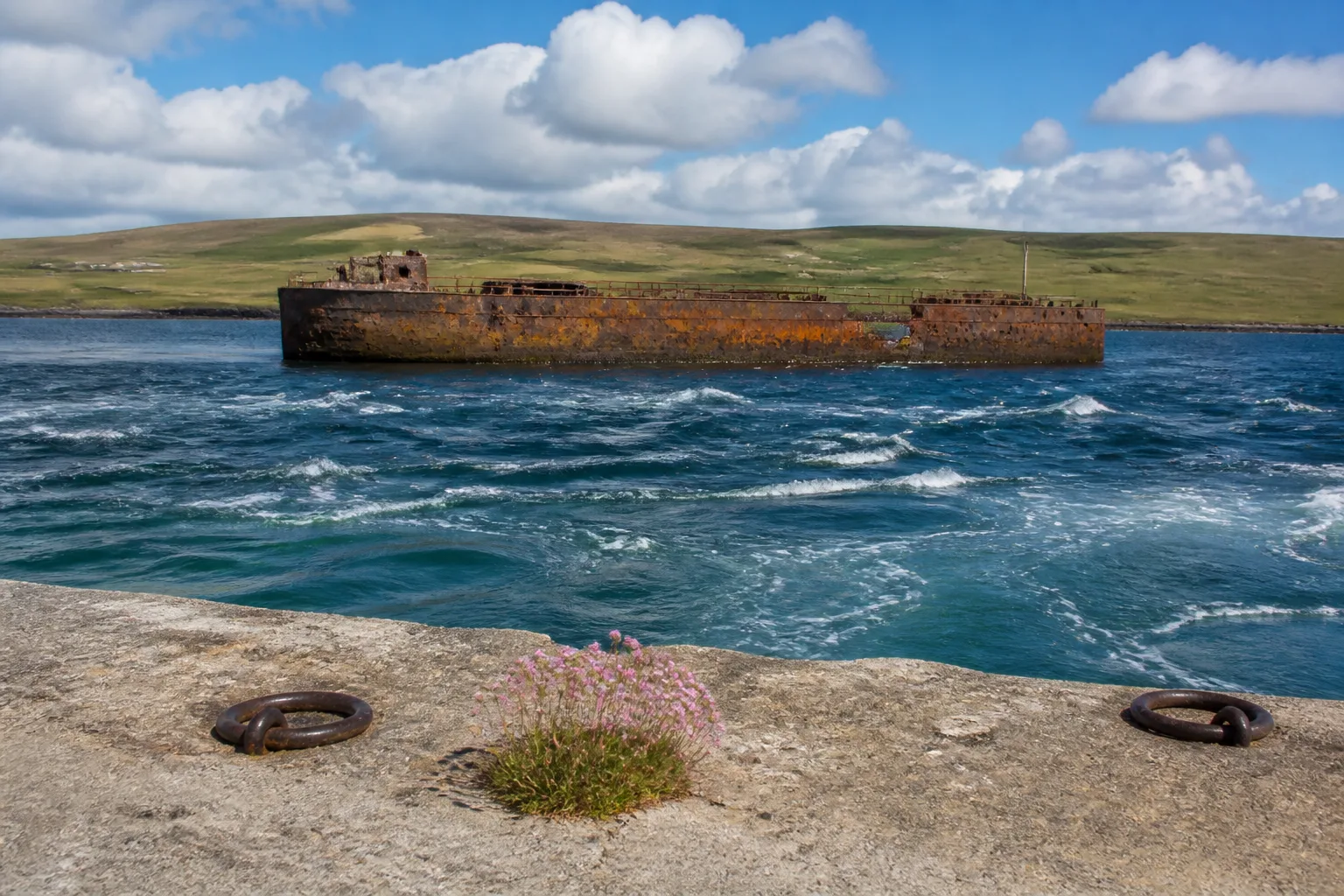 Rusted hull of one of the WWII blockships breaking the surface in the tidal blue water of Burra Sound, Orkney, with the green hills of Mainland in the background under broken summer cloud