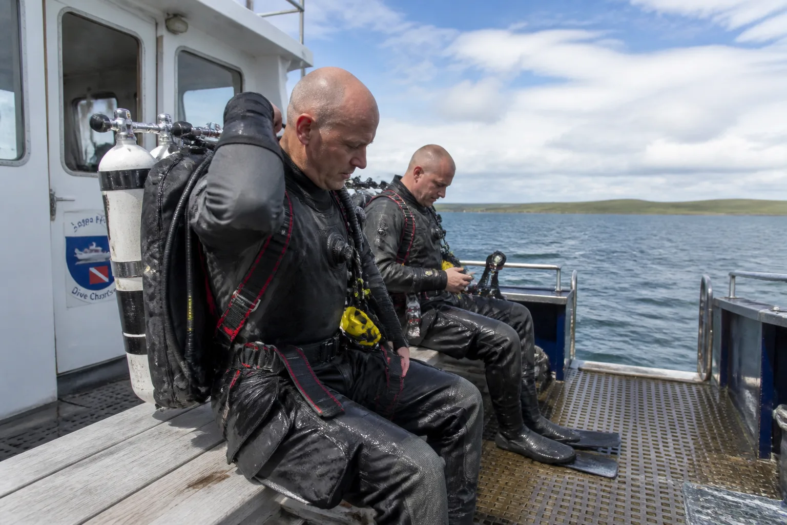 Two technical divers in grey neoprene drysuits kitting up with twin-cylinder backplate-and-wing rigs on the deck of a Scapa Flow charter boat at slack water, with the green coast of Hoy in the background