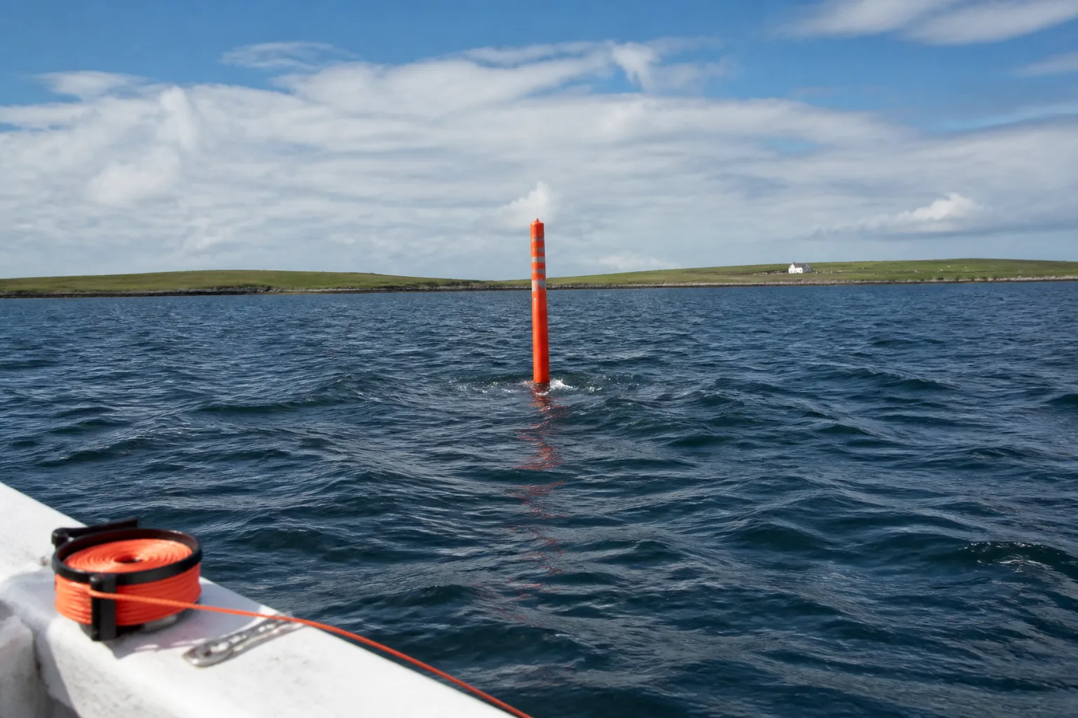 Orange surface marker buoy bobbing on the choppy blue water of Scapa Flow at slack water with the low green island of Cava in the background under a partly cloudy summer sky, photographed from the deck of a dive boat