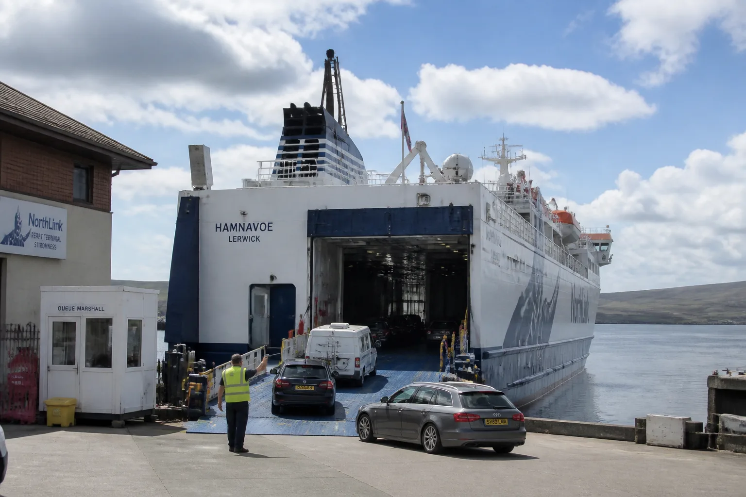 NorthLink ferry MV Hamnavoe berthed at Stromness ferry terminal on a bright partly-cloudy summer afternoon with three cars and a campervan driving slowly up the open vehicle loading ramp, harbour water calm with reflections of broken summer cloud, the hilly outline of Hoy faintly visible in the distance