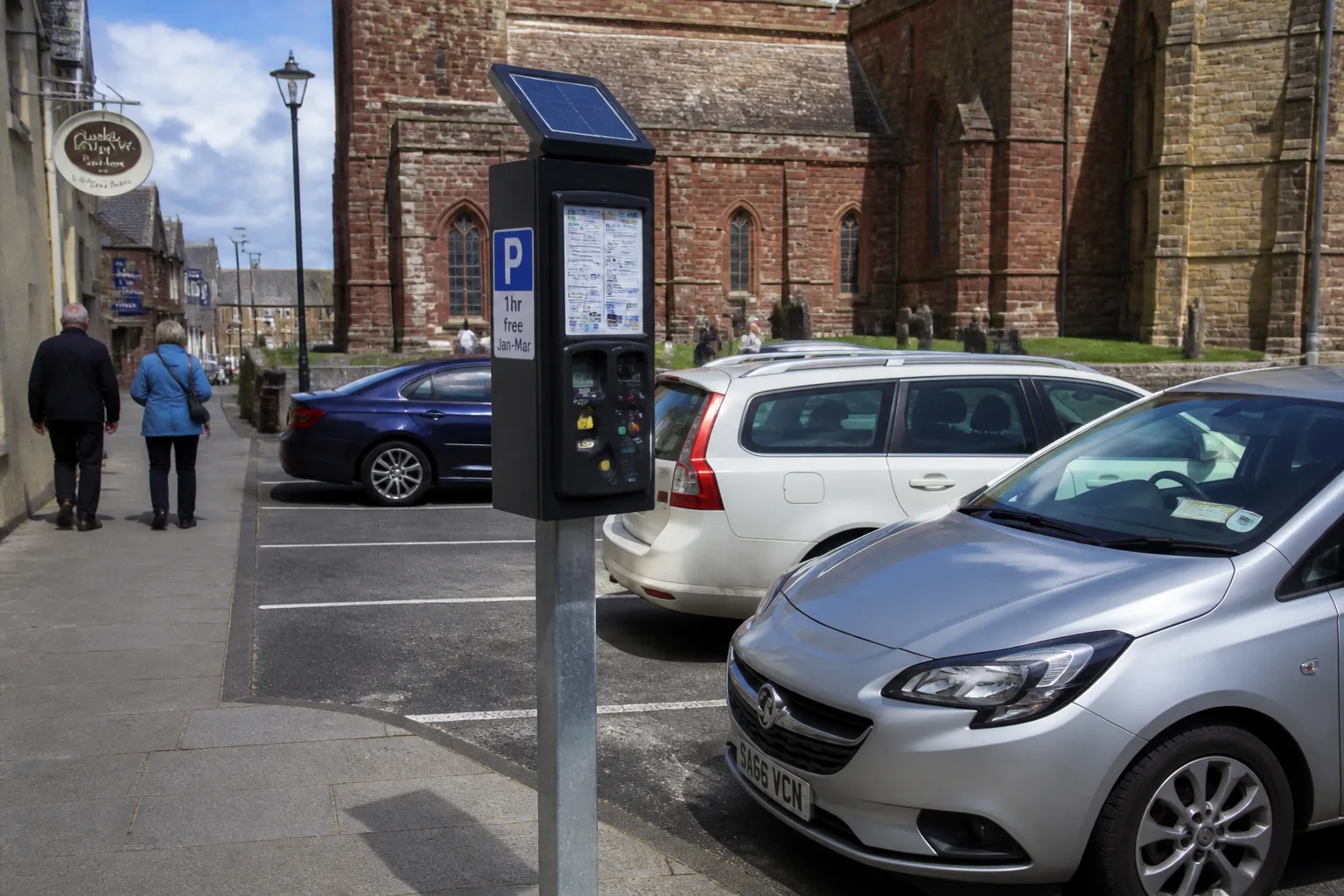 Cars parked on Broad Street in central Kirkwall on a bright partly-cloudy summer afternoon, St Magnus Cathedral red and yellow sandstone tower in the background, a pay-and-display machine on a metal post at the kerb with a small free January-March sticker, locals walking past with faces away from camera