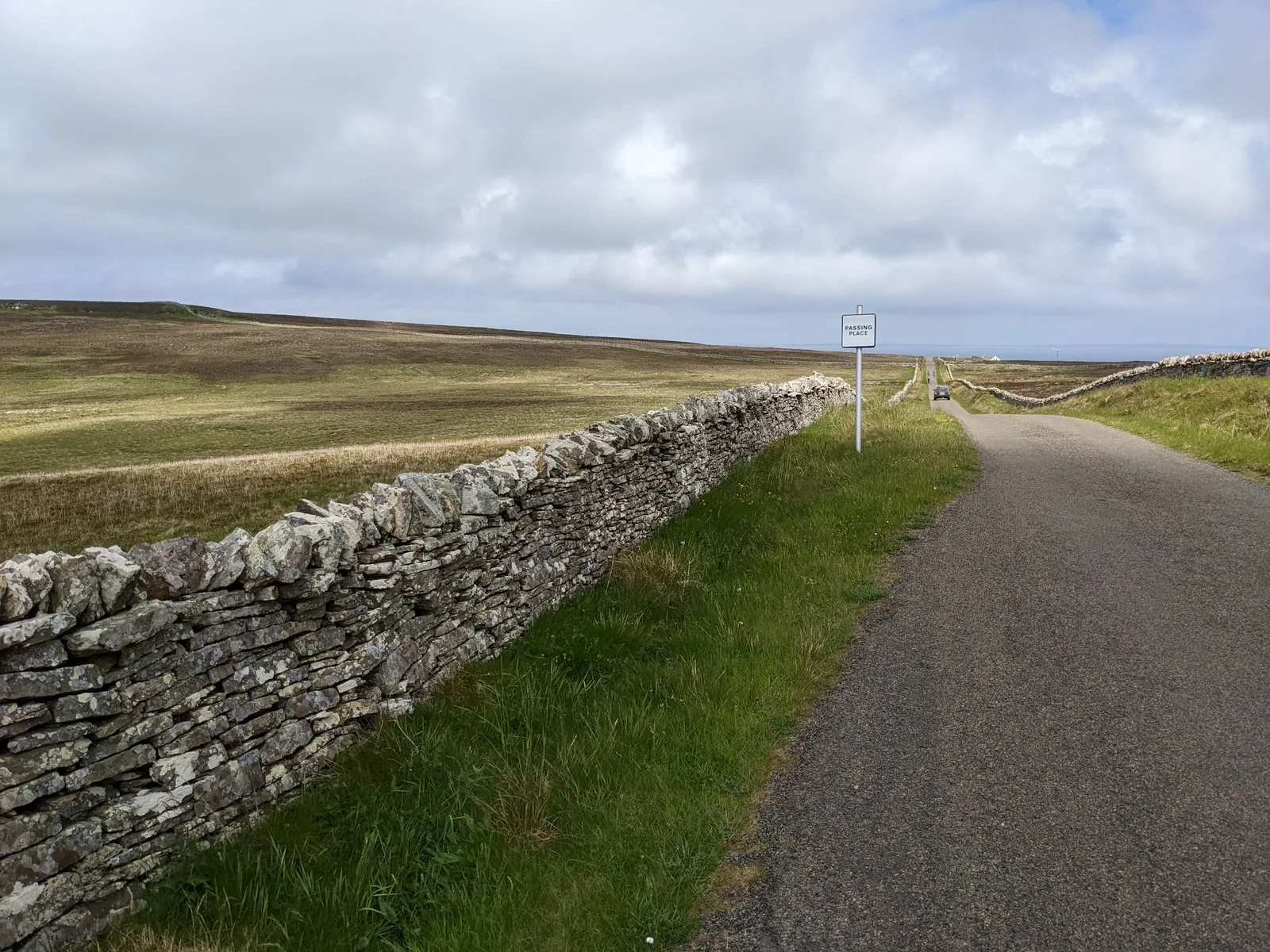 A single-track road on Orkney Mainland beside a drystone dyke, with a small square Passing Place sign at a widened section