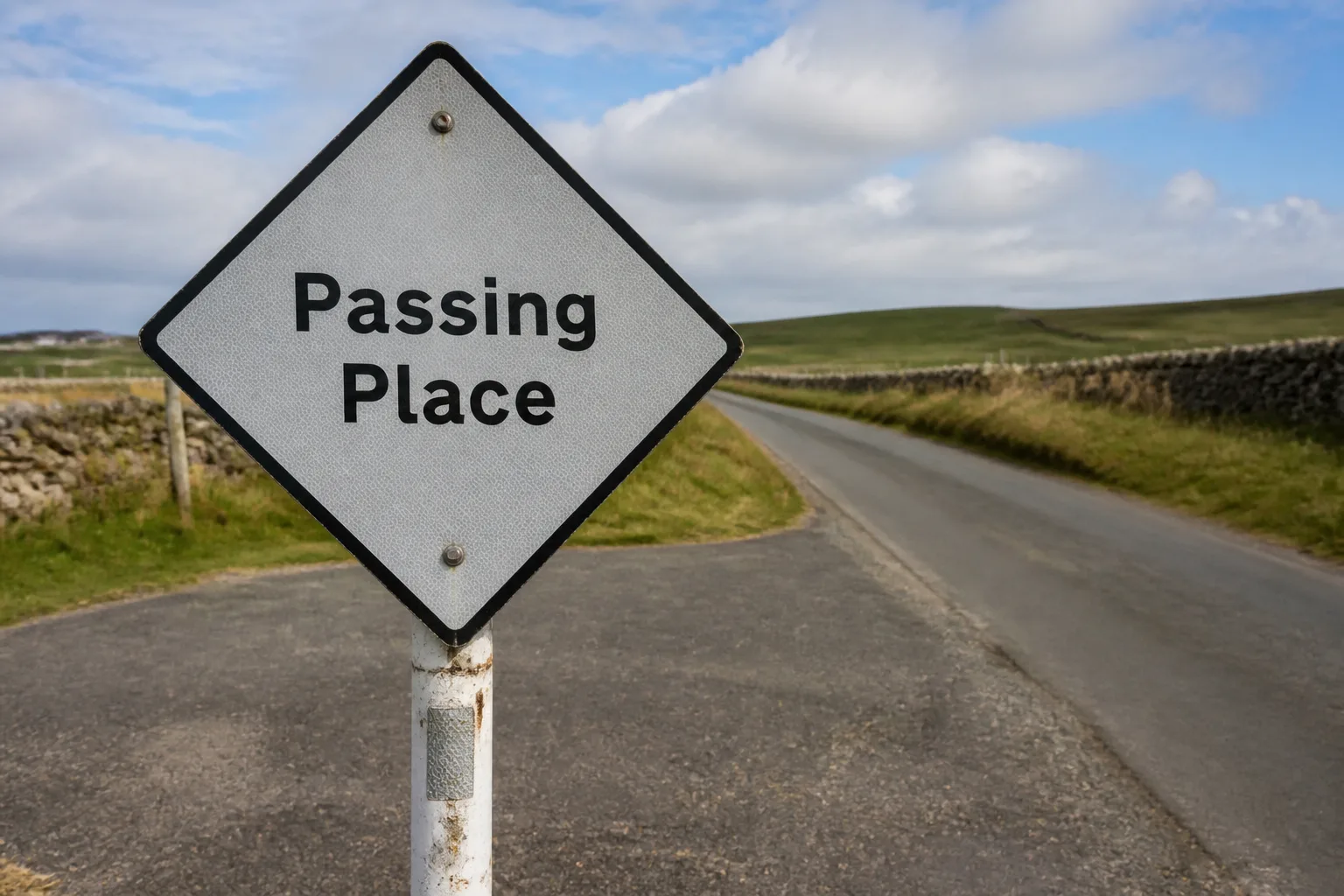 Tight close-up of a white diamond Passing Place sign on a short post at the edge of a single-track road on Orkney mainland, with the tarmac lay-by and a narrow road curving into soft-focus green farmland beyond on a bright partly-cloudy summer afternoon