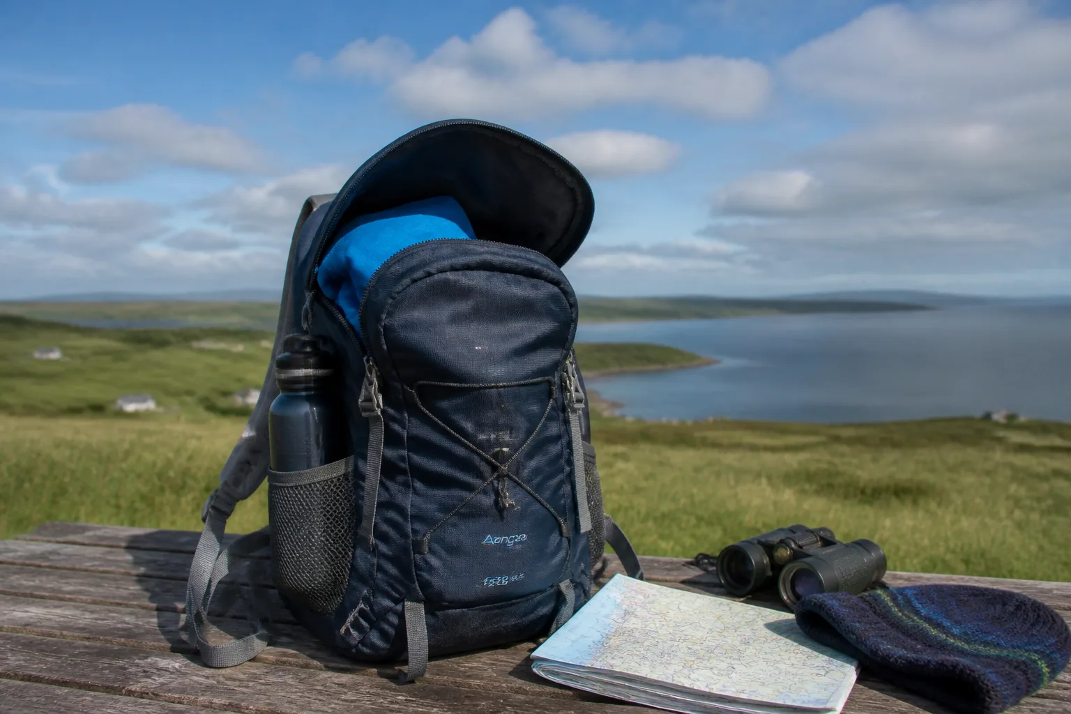 A 25-30 litre day backpack packed and open on a wooden bench at an Orkney viewpoint with a rolled waterproof, water bottle, binoculars, paper map and wool hat