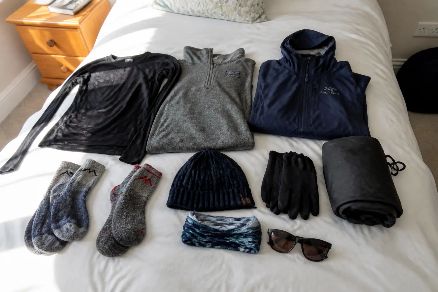 Flat lay of Orkney travel layers on a guest-house bed: merino base layer, grey fleece, navy waterproof shell, wool walking socks, beanie hat, gloves, buff and sunglasses