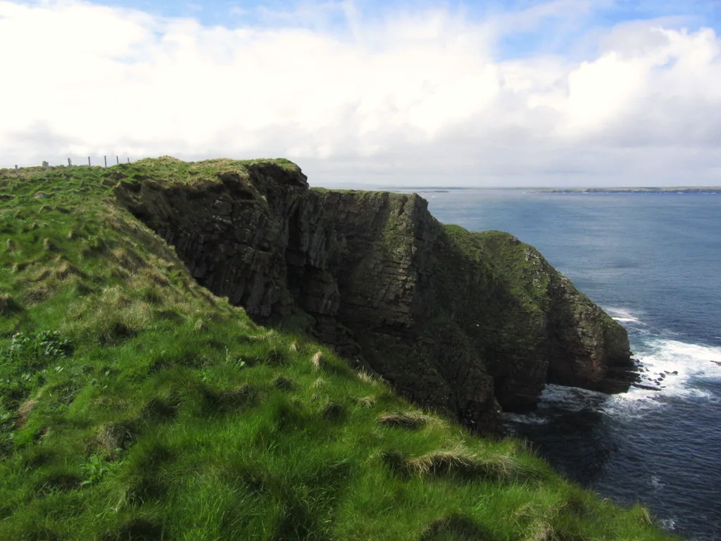 Sea cliffs at Red Head on the north coast of Eday, Orkney — layered Old Red Sandstone dropping to the deep blue Atlantic, with a grassy clifftop in the foreground.