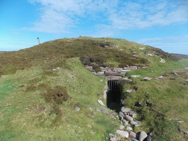The Vinquoy chambered cairn on Eday, Orkney — a grassy Neolithic mound on Vinquoy Hill with a gated stone entrance set below the summit cairn marker, under a bright blue summer sky.