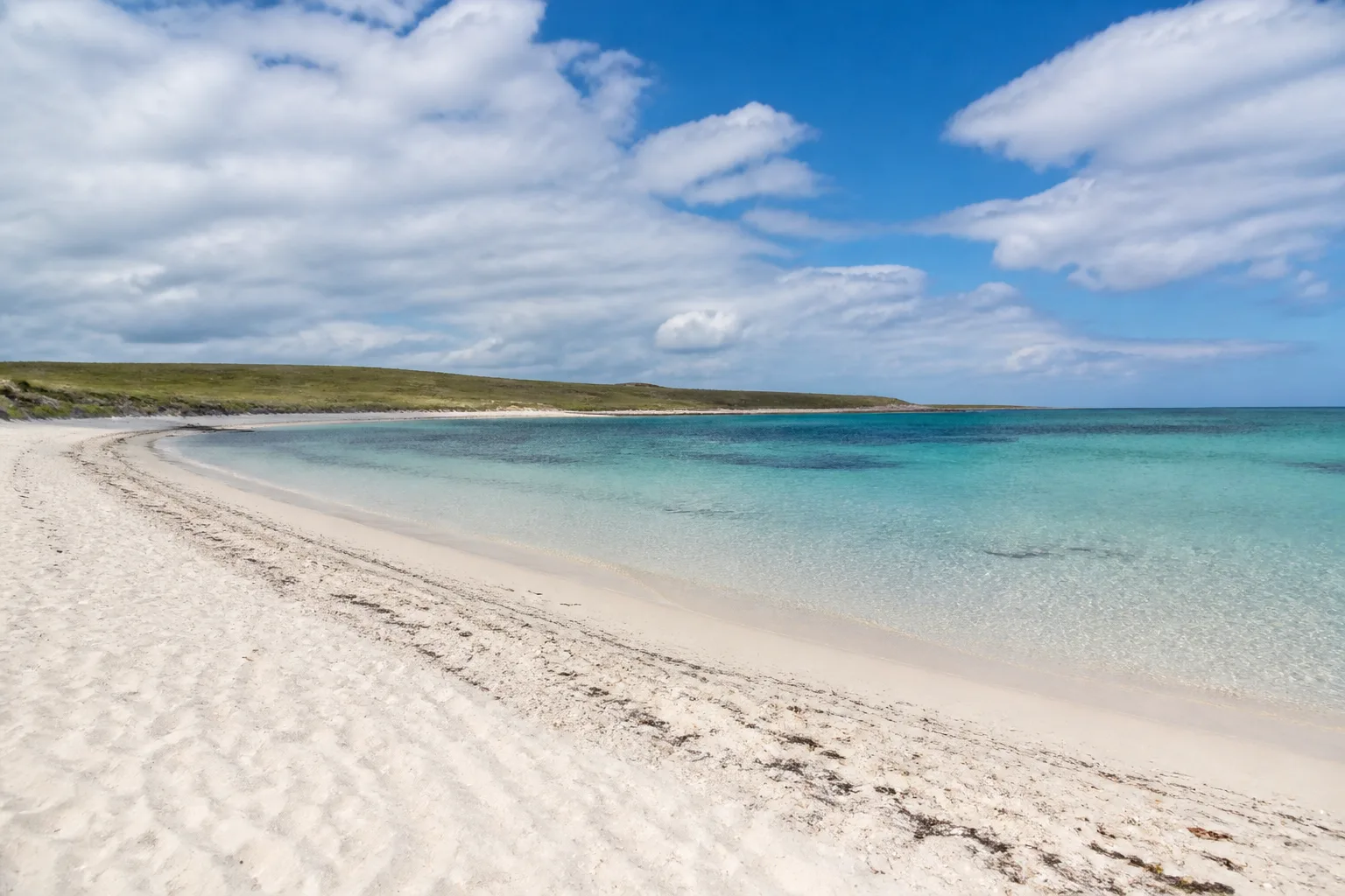 A wide empty pale shell-sand beach on North Ronaldsay with clear turquoise shallow water, low green dunes behind and a scattering of seaweed along the tideline, bright summer day with broken cloud