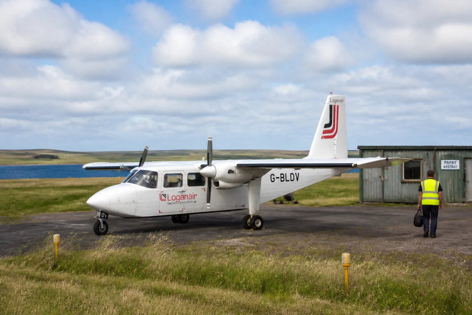Small white Loganair Britten-Norman Islander twin-prop aircraft parked on the short Papa Westray airstrip on a bright partly-cloudy summer day, with the green Papay farmland and blue strait to Westray visible in the background