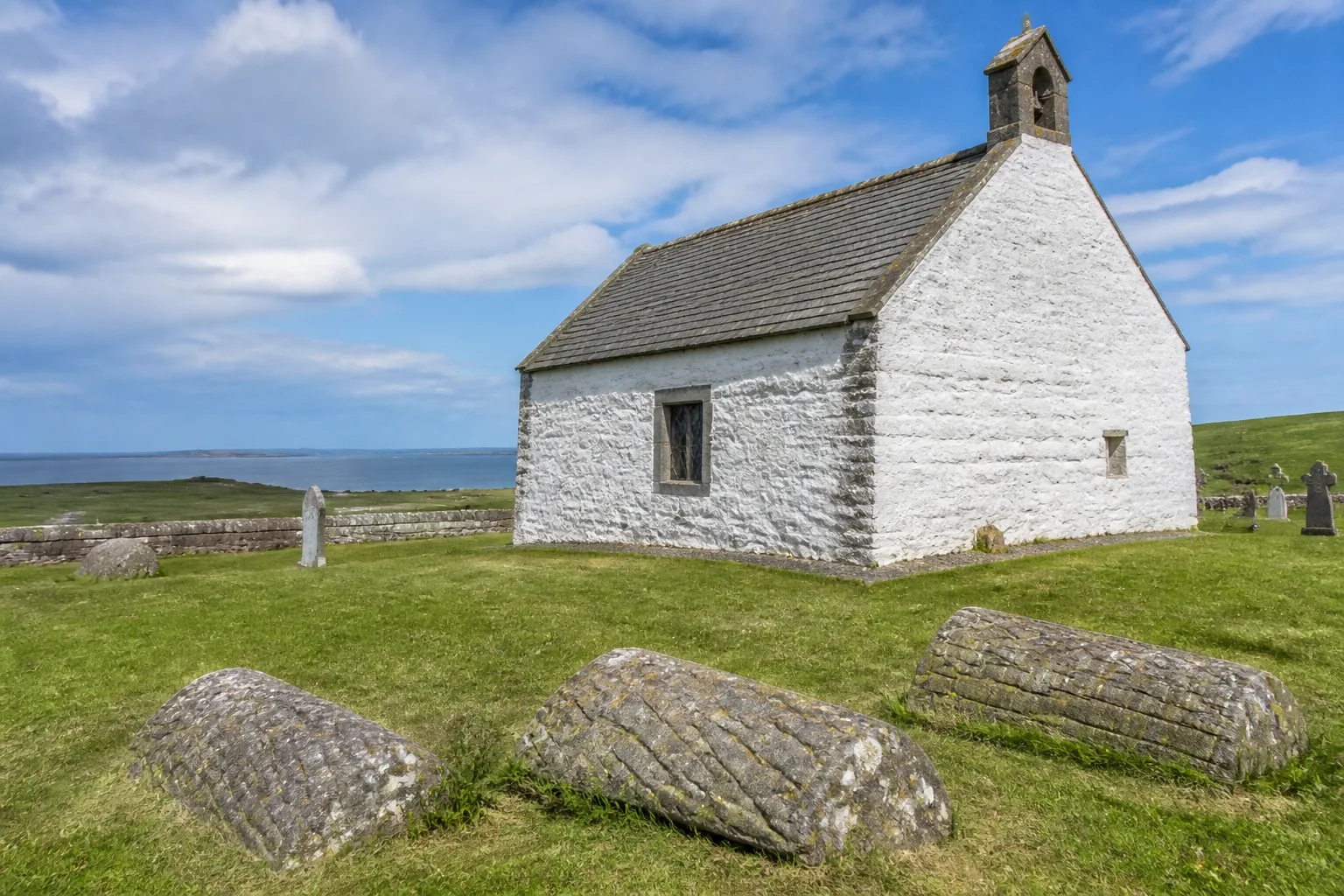 Small medieval St Boniface Kirk on Papa Westray Orkney with three weathered Norse-era hogback gravestones in the foreground grass and the open blue sea visible in the background under summer broken cloud