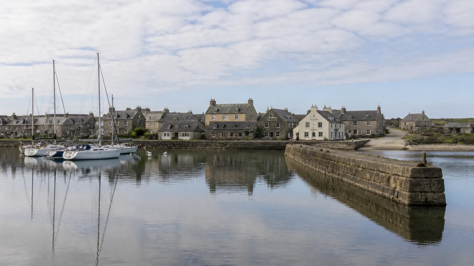 A wide photo of Balfour village on Shapinsay from across the pier — the symmetrical 19th-century planned village around the salt store and gatehouse, the stone Dishan Tower at the shore edge, the long stone pier in the foreground with MV Shapinsay tied up, neat estate-style terraces with grey slate roofs running back from the harbour, the green land rising to a treeline that screens Balfour Castle.