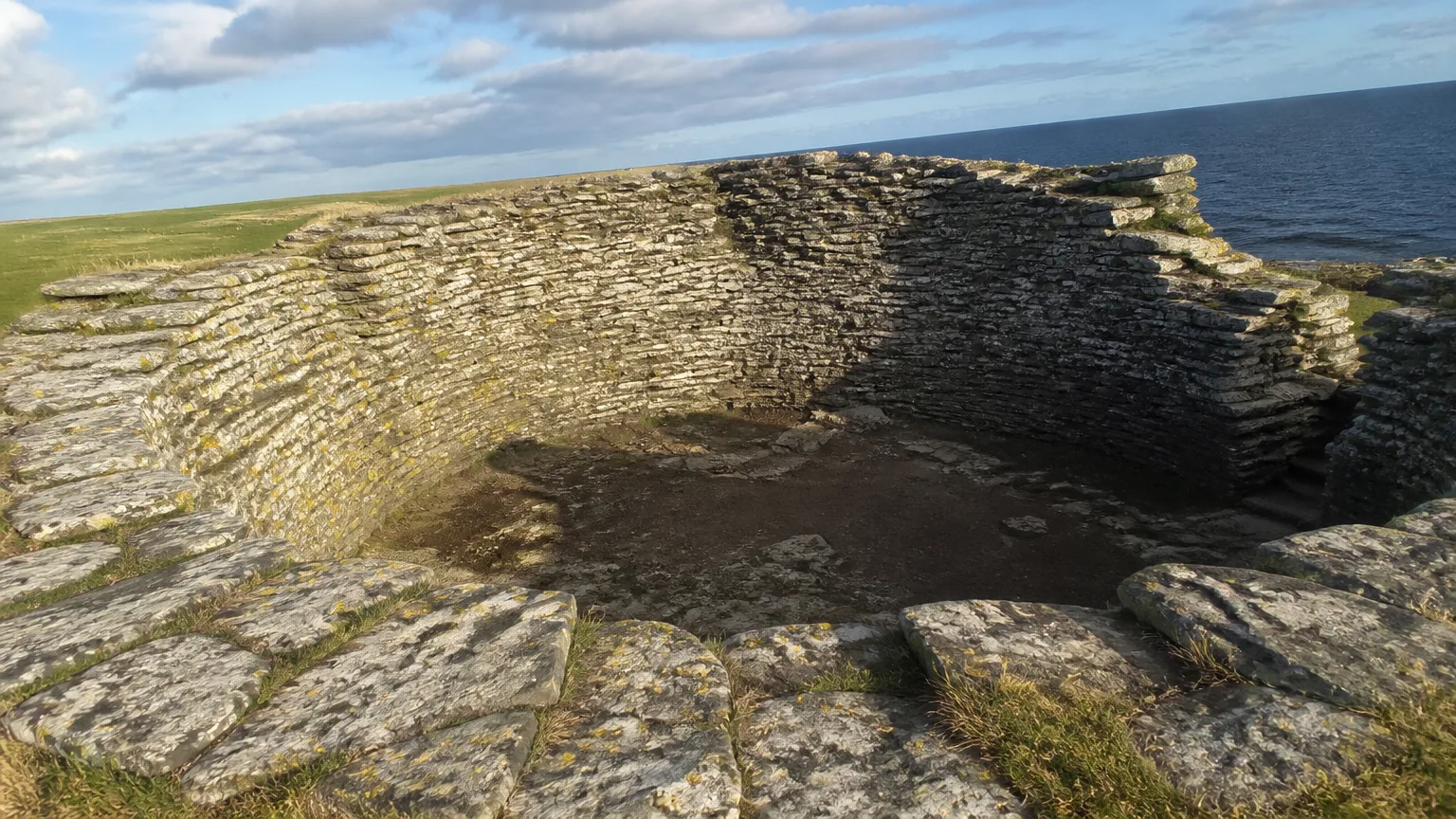 Burroughston Broch on the northeast coast of Shapinsay — Iron Age drystone walls standing to roughly three metres in a low grass-covered mound on a flat headland with the sea beyond, the cleared circular interior in the foreground showing courses of carefully-fitted flat stones, the entrance passage on the left, the dark rocks of the seal haul-out at the shore edge, sheep grazing under broken Orkney cloud.