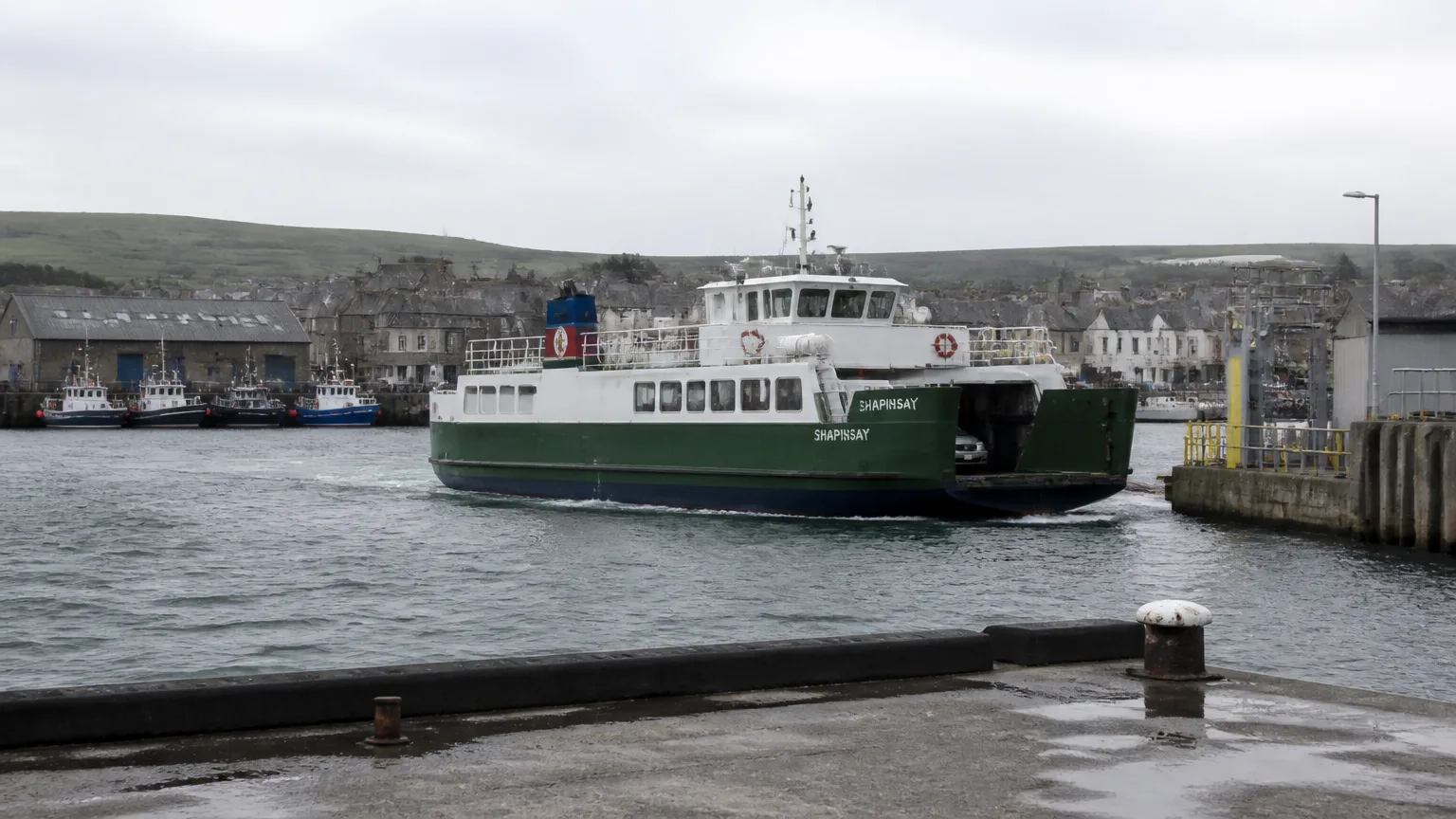 MV Shapinsay tied up at Kirkwall harbour pier — the small green-and-white Ro-Ro ferry with its open vehicle deck at the stern and a single passenger deck above, gangway down with a few foot passengers walking aboard with bicycles, masts of Kirkwall fishing boats and the spire of St Magnus Cathedral behind the ferry, calm grey-blue water in the foreground, soft overcast light.