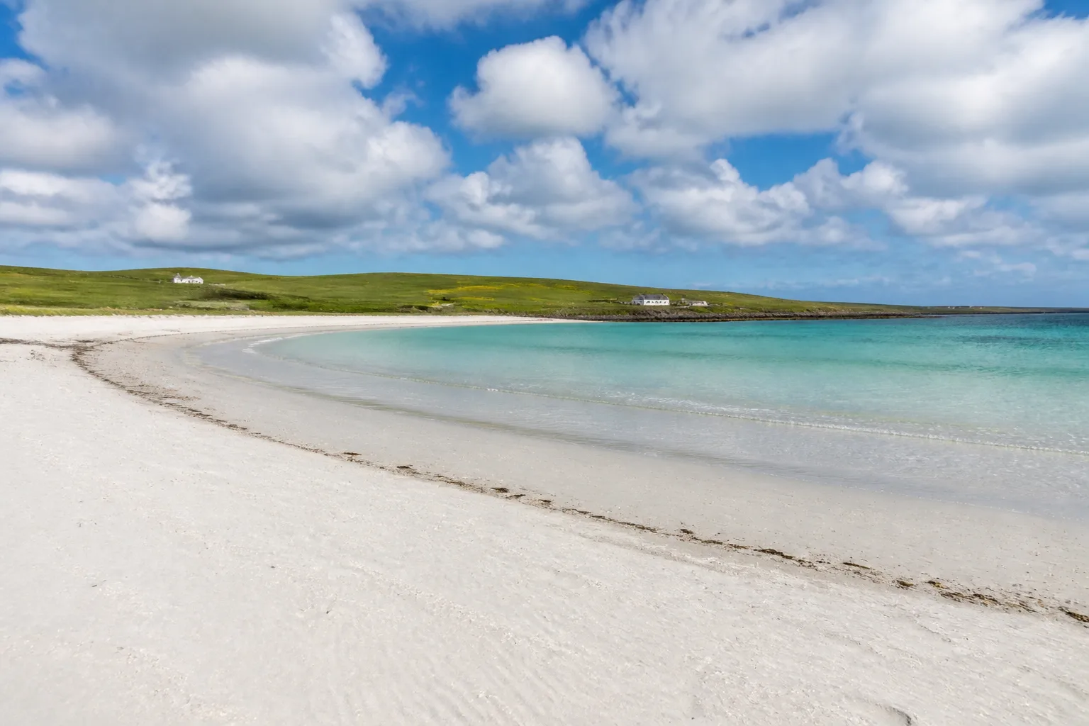 The wide empty white shell-sand beach of St Catherine's Bay on Stronsay Orkney curving around a sheltered bay with clear turquoise shallow water, low green farmland and a couple of distant white farmhouses behind, on a bright partly-cloudy summer day
