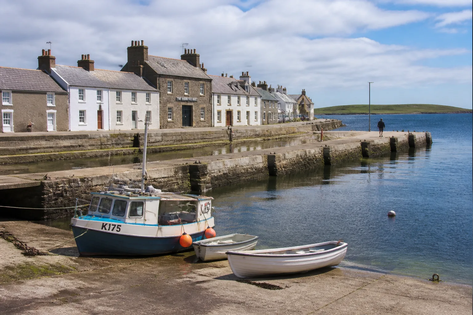 Whitehall village harbour on Stronsay Orkney — a row of low white-harled and grey-stone houses along a stone harbour front, with a small fishing boat and dinghies at a slipway in the foreground and a calm blue bay behind, on a bright partly-cloudy summer day