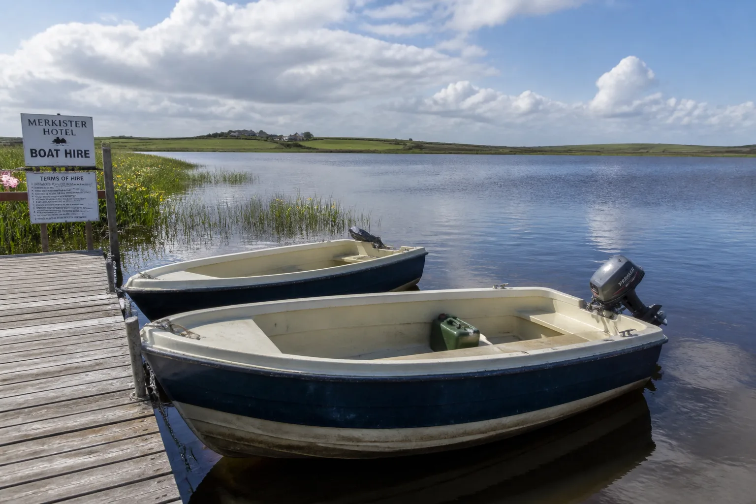 Two traditional fibreglass rowing boats moored at a small wooden jetty on the Loch of Harray on a bright partly-cloudy summer morning, weathered cream-and-blue hulls with outboard, summer reeds and yellow flag iris on the bank