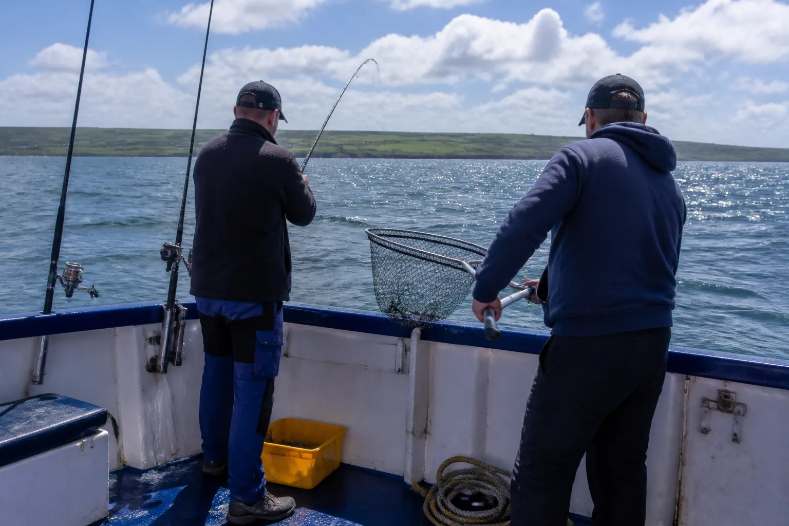 View from the open back deck of a small white-and-blue sea-angling charter boat on Scapa Flow on a bright partly-cloudy summer day with two casual anglers at the stern rail, one with rod bent into a fish, the other holding a landing net