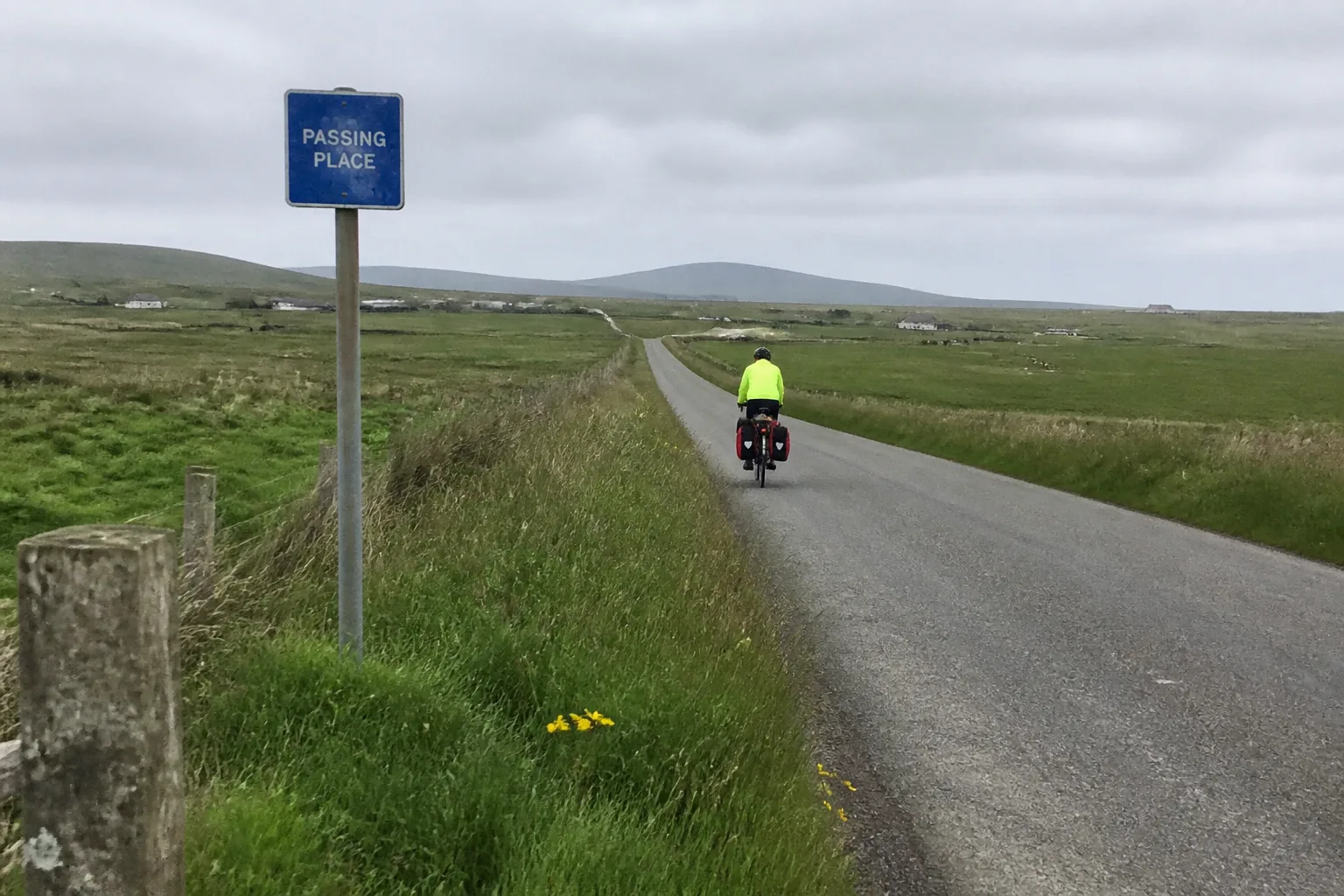 A single touring cyclist riding away from the camera along an empty single-track tarmac road across the West Mainland of Orkney, panniers loaded, wearing a yellow high-vis cycling jacket, with green farmland on either side and a passing-place sign on the left