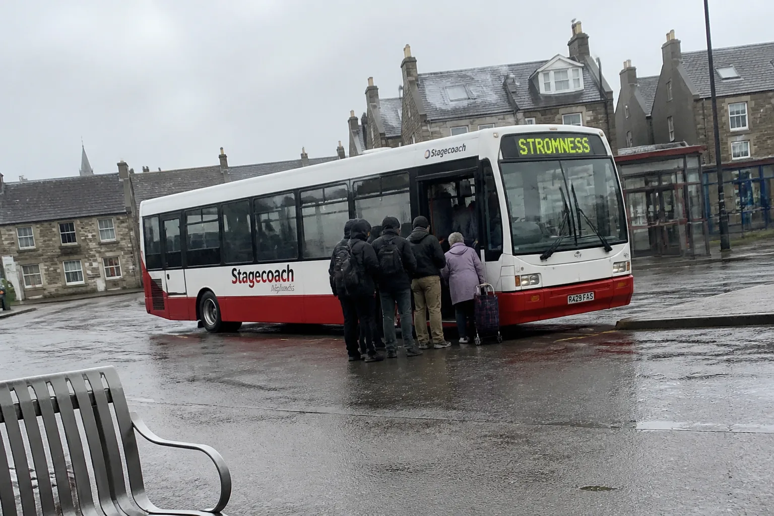 A white-and-red Stagecoach Highlands single-decker bus parked at Kirkwall bus station in Orkney with the destination display reading STROMNESS, a few passengers in waterproof jackets queuing to board including an older woman with a wheeled shopping bag, on an overcast morning