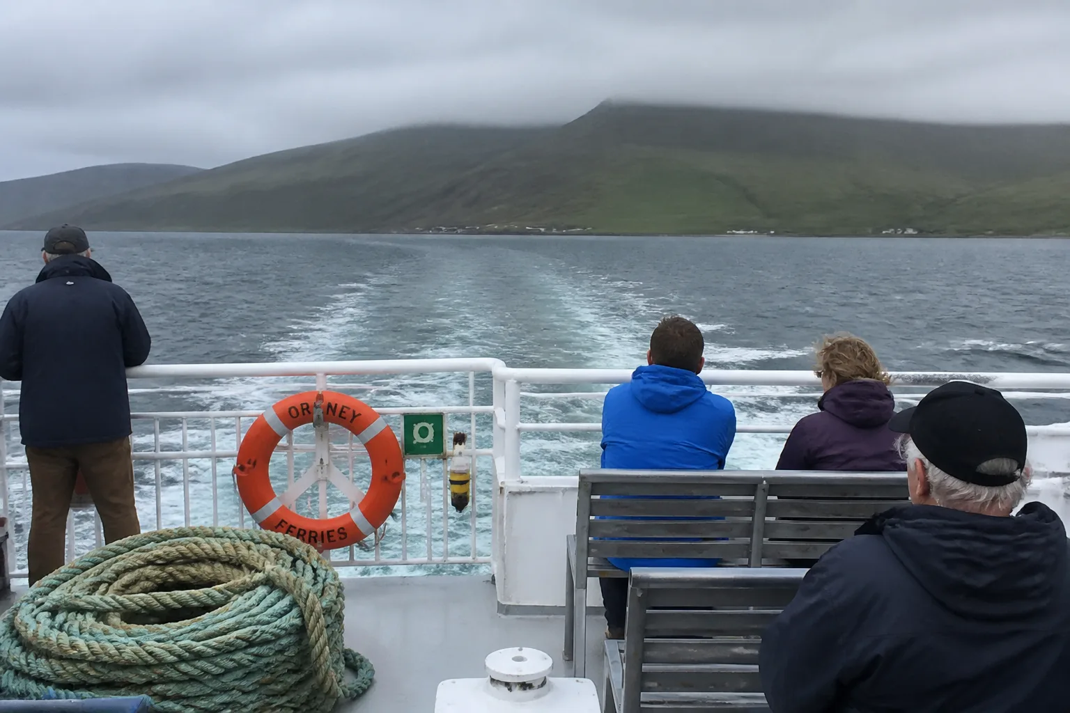 The open passenger deck of a small Orkney Ferries roll-on/roll-off ferry between Houton and Lyness on Hoy, with white-painted deck and metal bench seats, a few passengers in waterproof jackets at the rail watching the wake, and the green hills of Hoy emerging from low cloud in the distance