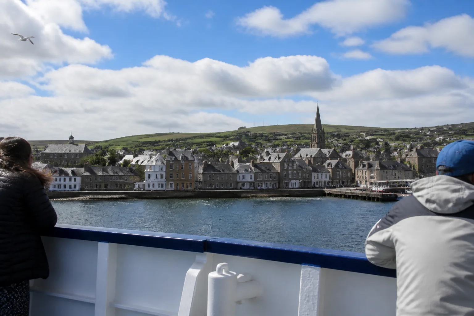 View from the deck of NorthLink ferry MV Hamnavoe approaching Stromness harbour, Orkney, on a bright partly-cloudy summer morning — slate-roofed sandstone houses lining the waterfront with a church spire above, low green hills of West Mainland in the background, white-painted ferry rail in the foreground