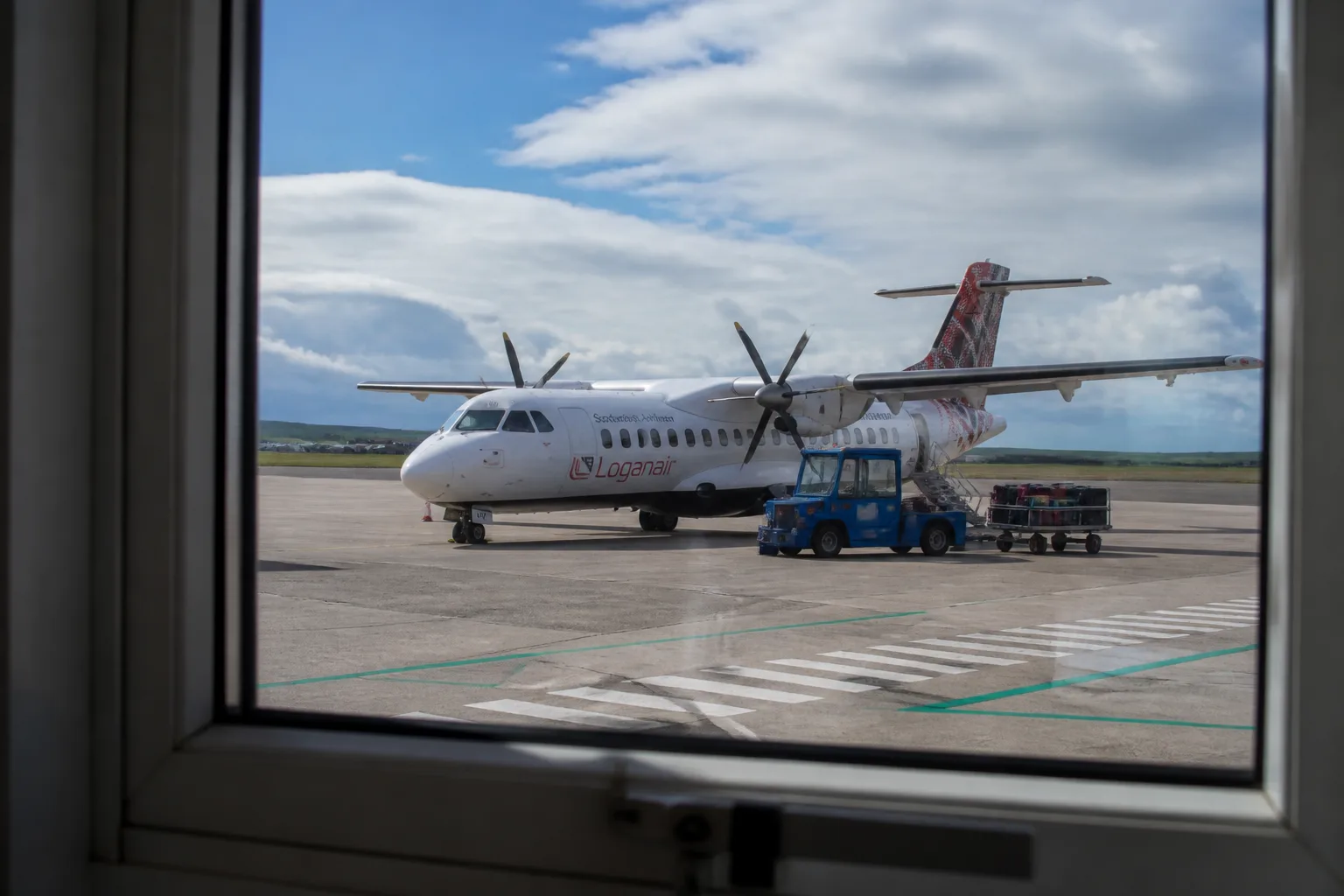View through Kirkwall Airport terminal windows onto the apron — a Loganair ATR 42 turboprop aircraft with white fuselage and red-and-grey Saltire tail livery parked on the tarmac in bright partly-cloudy summer light, ground-handling vehicle beside it