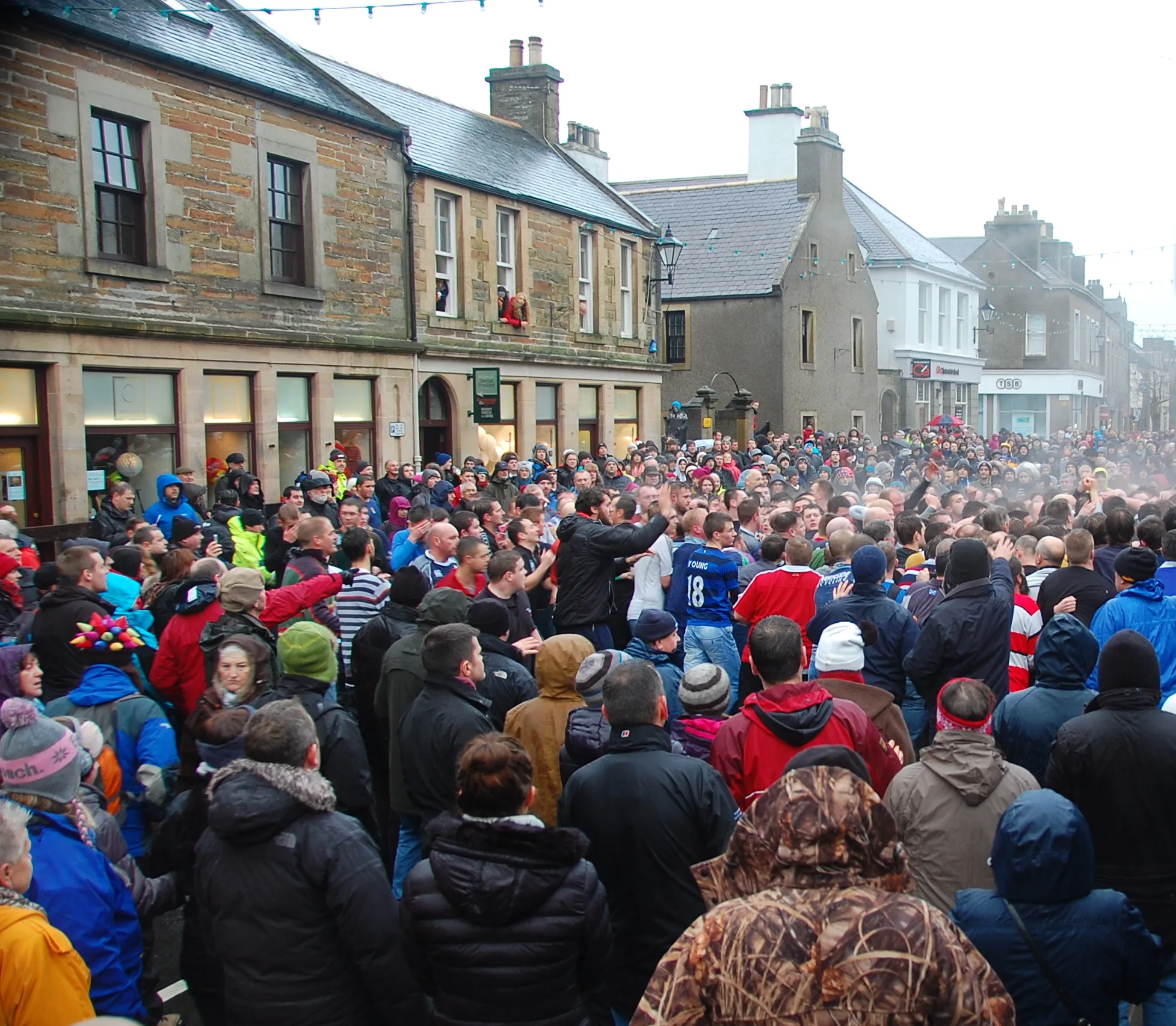 Men packed tightly against a stone wall and railing during the Kirkwall Ba', reaching up and climbing to escape the crush of the scrum on Broad Street