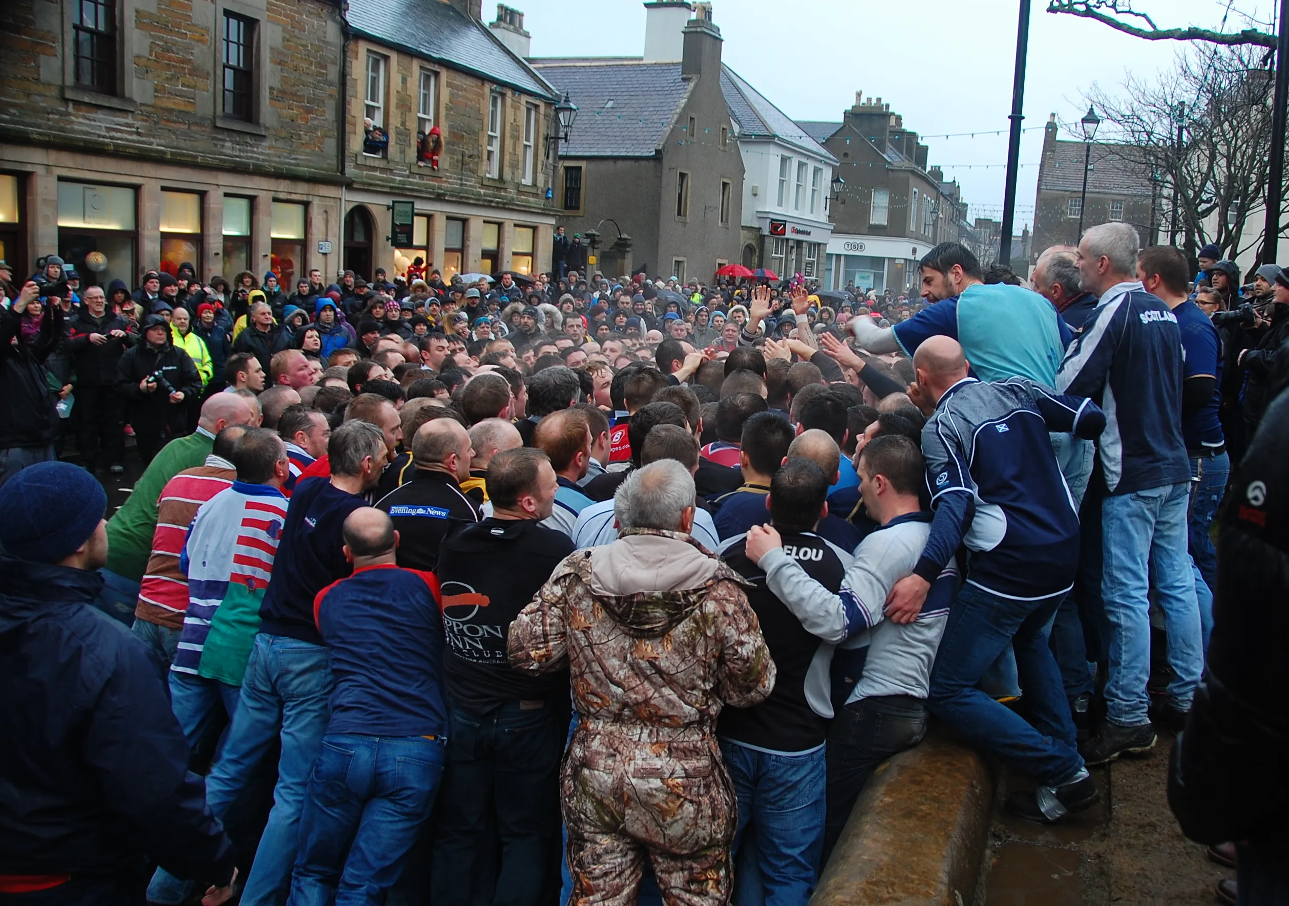 A dense scrum of men packed together on Broad Street, Kirkwall, in front of shopfronts with their windows boarded up for the Ba', with spectators watching from upstairs windows