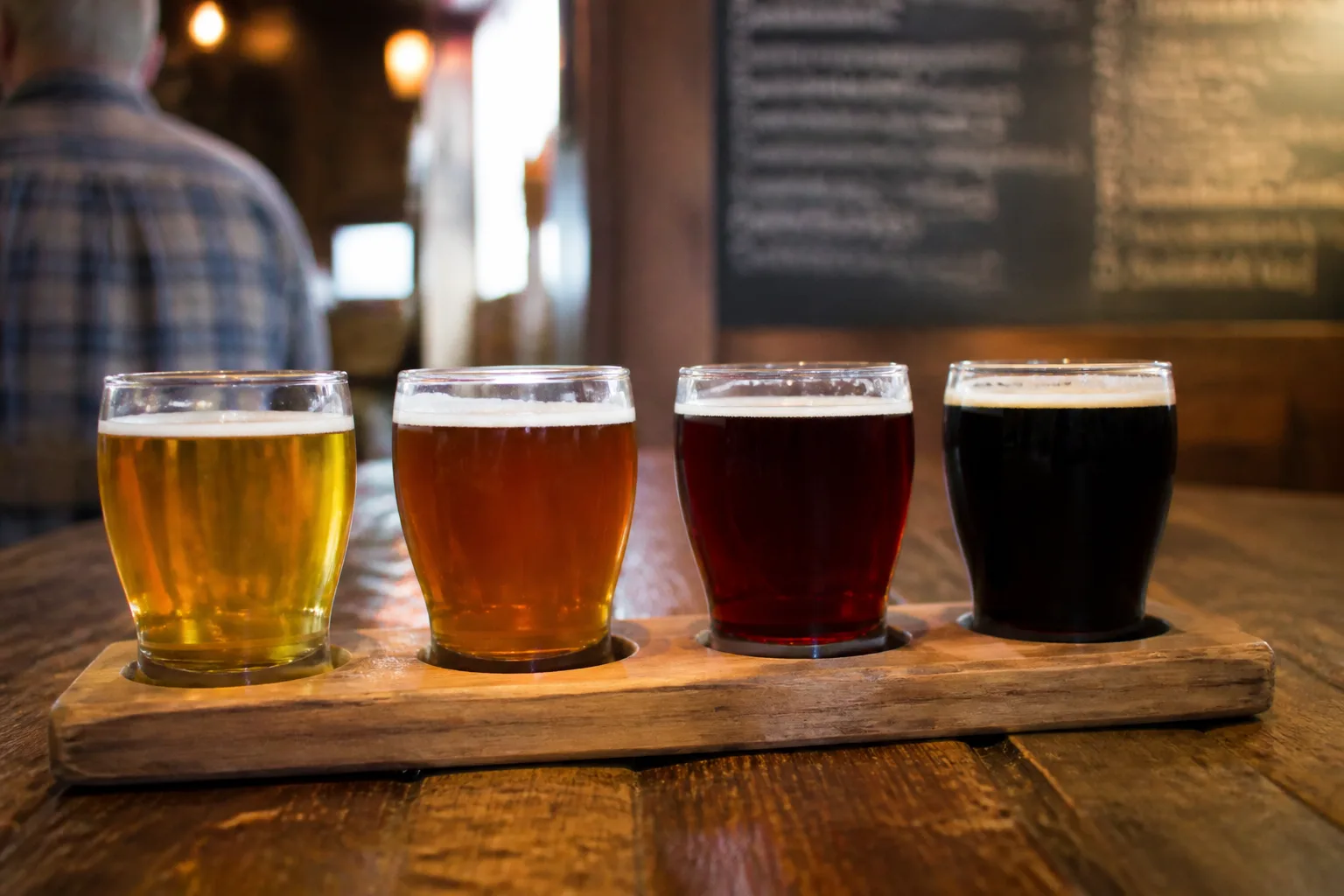 An amateur Canon DSLR photo of a tasting flight of four half-pint glasses holding Orkney ales of different colours, from pale gold to dark ruby to near-black, arranged on a rustic wooden pub table with a hand-written chalkboard menu blurred in the background