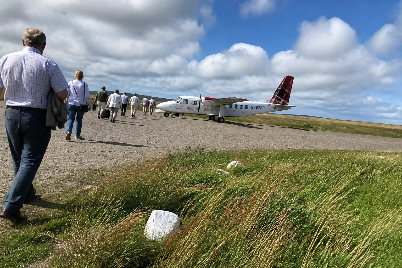 Small white twin-propeller Loganair Britten-Norman Islander aircraft sitting on the gravel-and-grass airstrip at Papa Westray, with passengers in waterproof jackets walking towards the plane under a heavy overcast sky